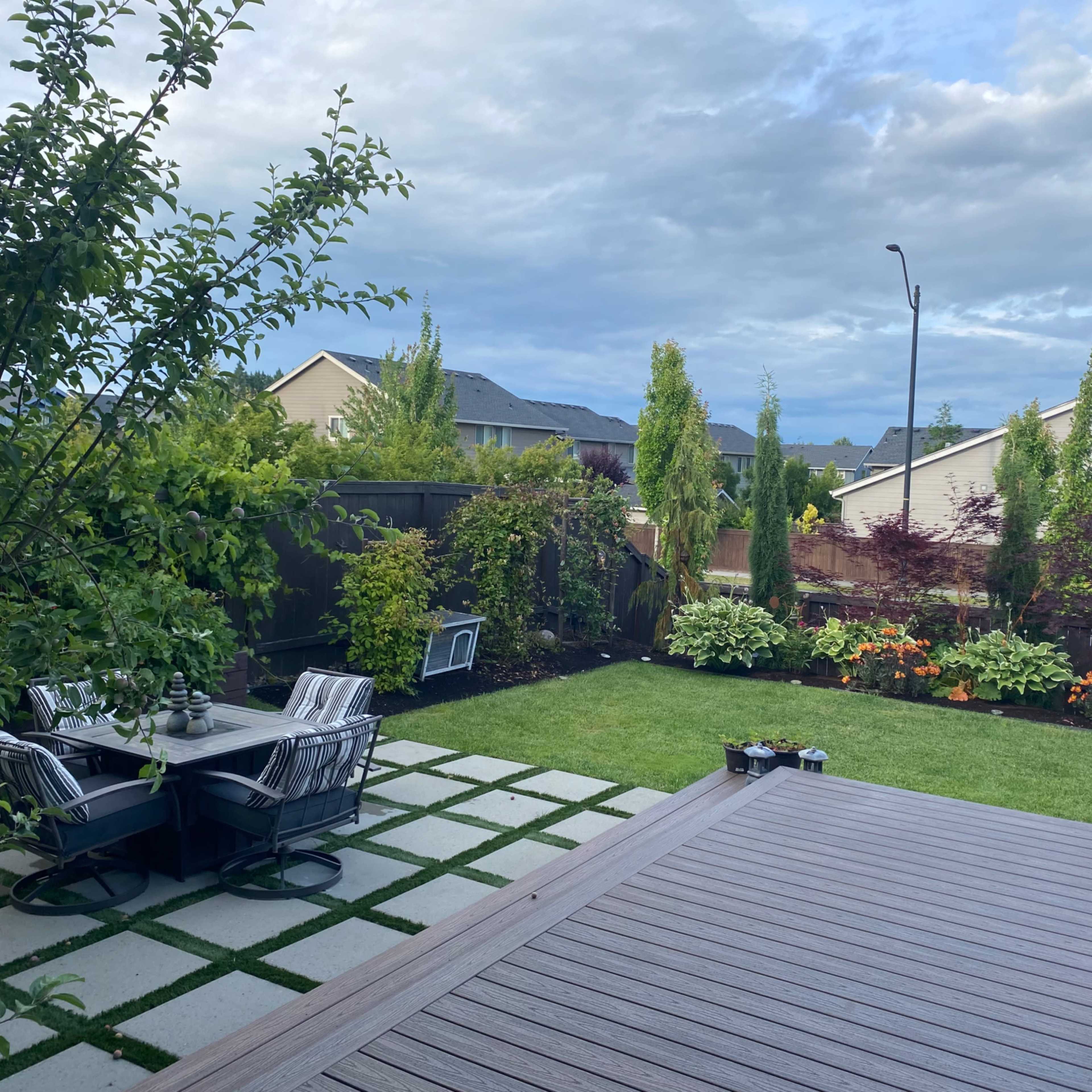 A backyard garden features a patio set on stone tiles, surrounded by neatly arranged flower beds and lush greenery under a cloudy sky.
