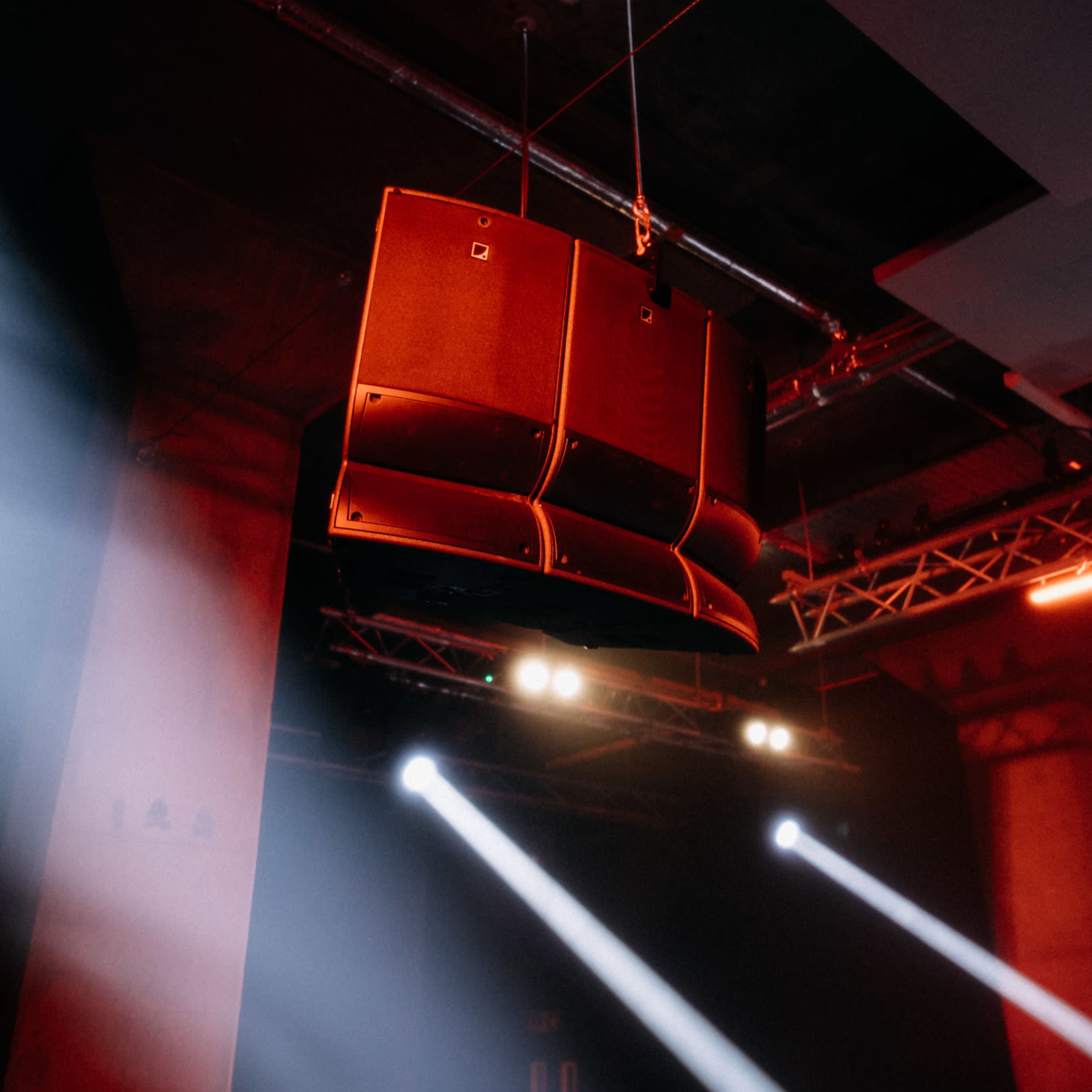 A set of large sound speakers is suspended from the ceiling in a dimly lit venue with beams of light shining through the smoke.