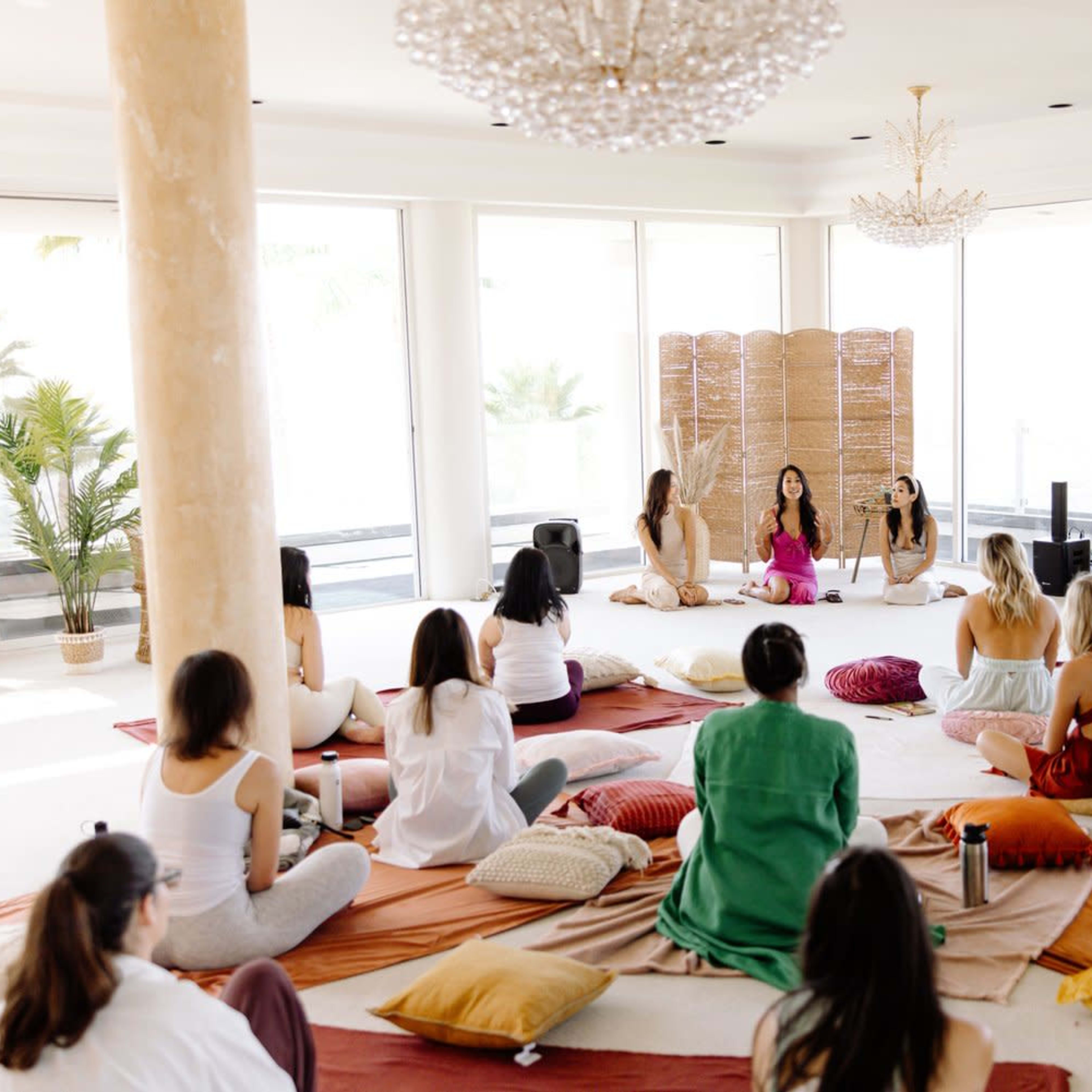 A group of people sits on floor cushions in a bright, open space while a few individuals lead a discussion in front of a bamboo screen.