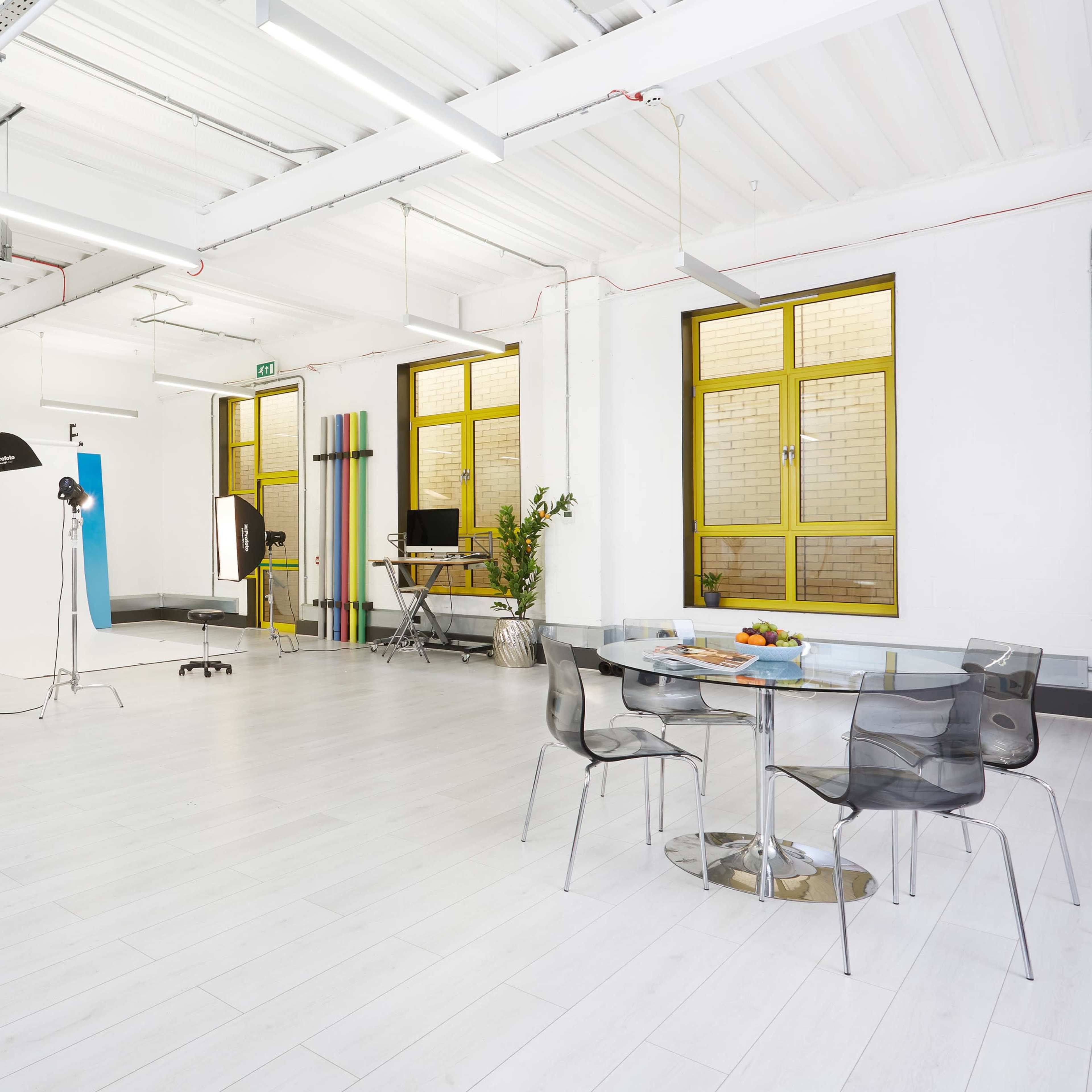 A spacious, well-lit studio features white walls, a gray sofa, and a glass dining table surrounded by transparent chairs, with a photography setup in one corner.