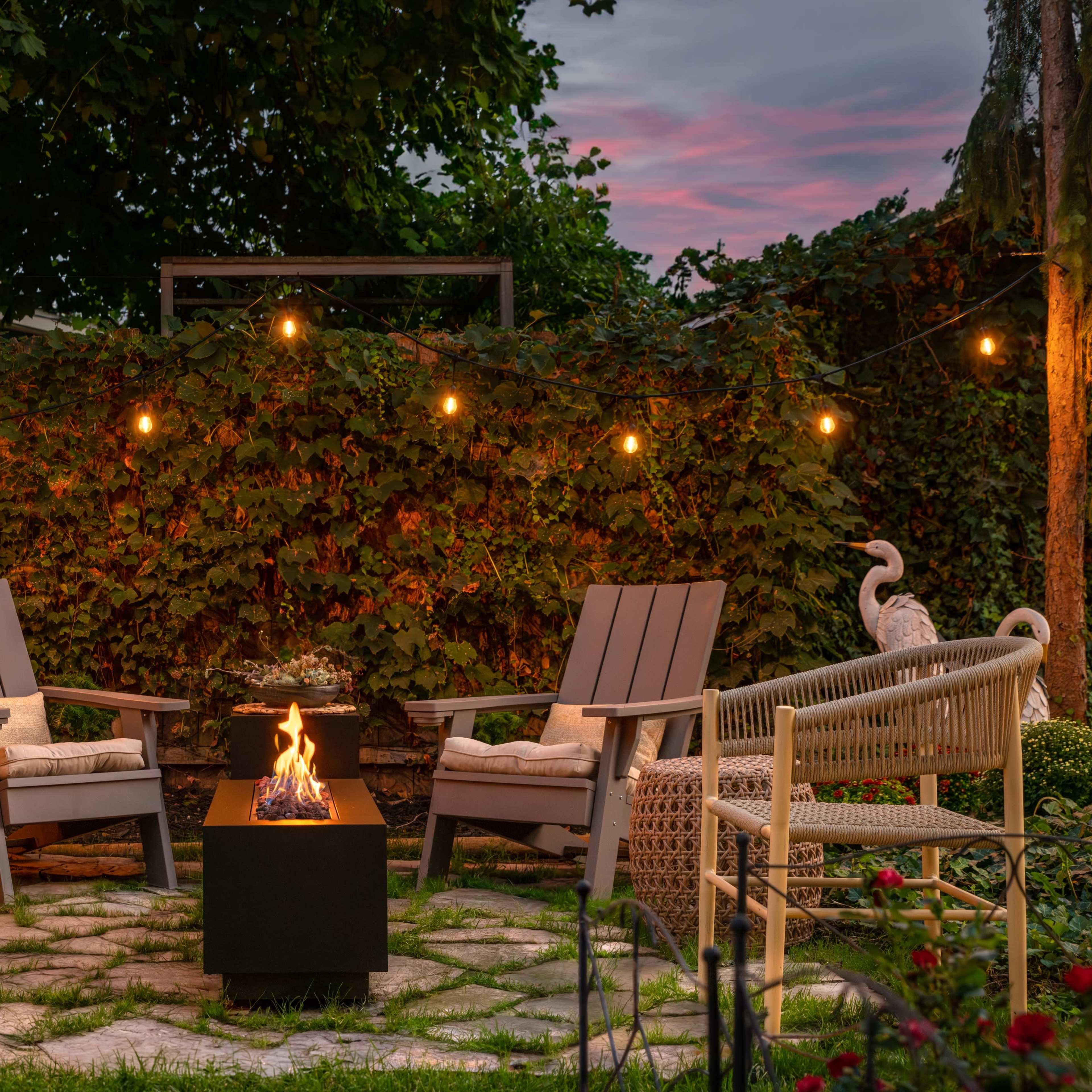 A cozy outdoor seating area features two chairs facing a fire pit surrounded by lush greenery and string lights.