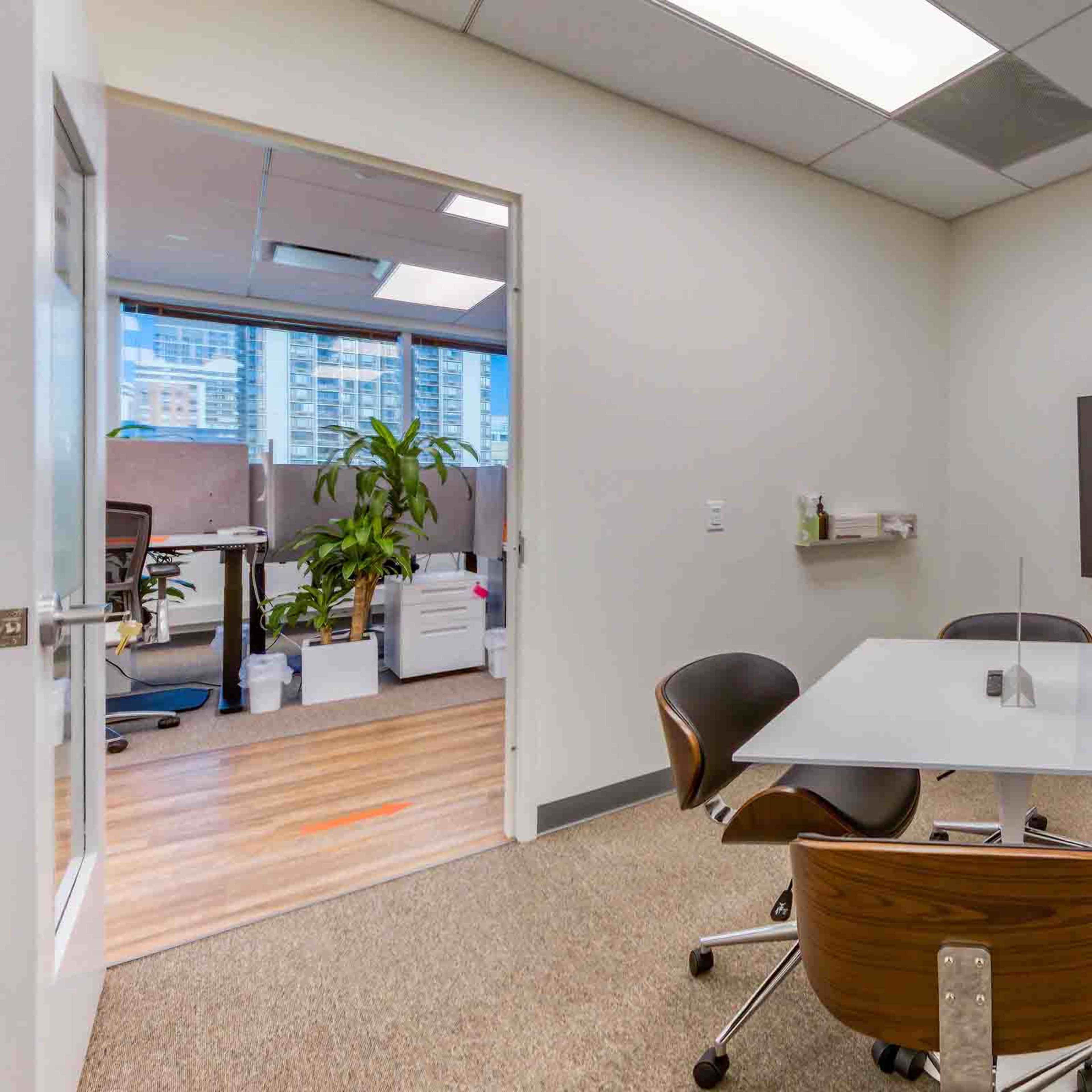 A small meeting room features a white table surrounded by four brown chairs, with a television mounted on the wall and an open office area visible through the doorway.