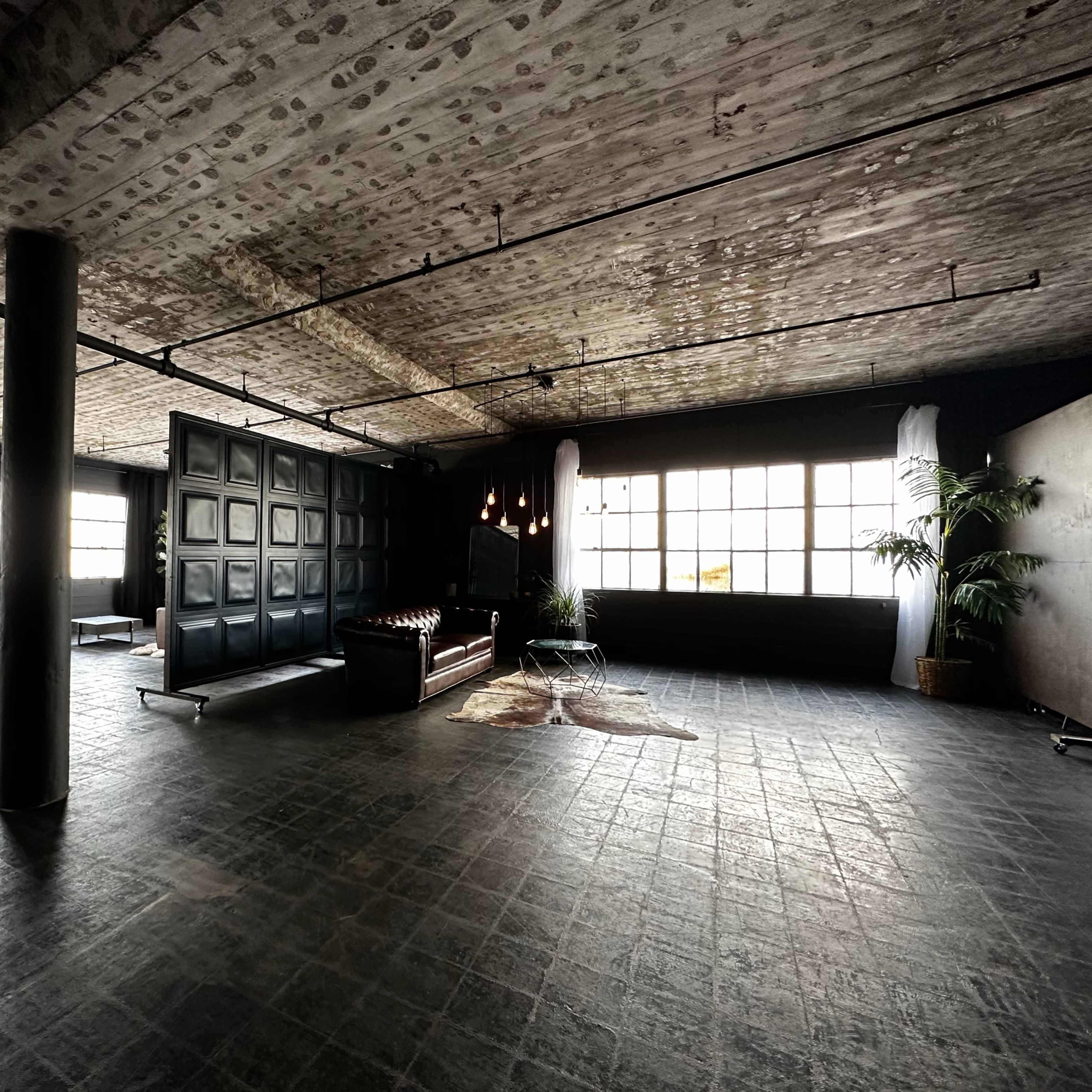 A spacious, industrial-style room with large windows, a leather sofa, and minimal furnishings.