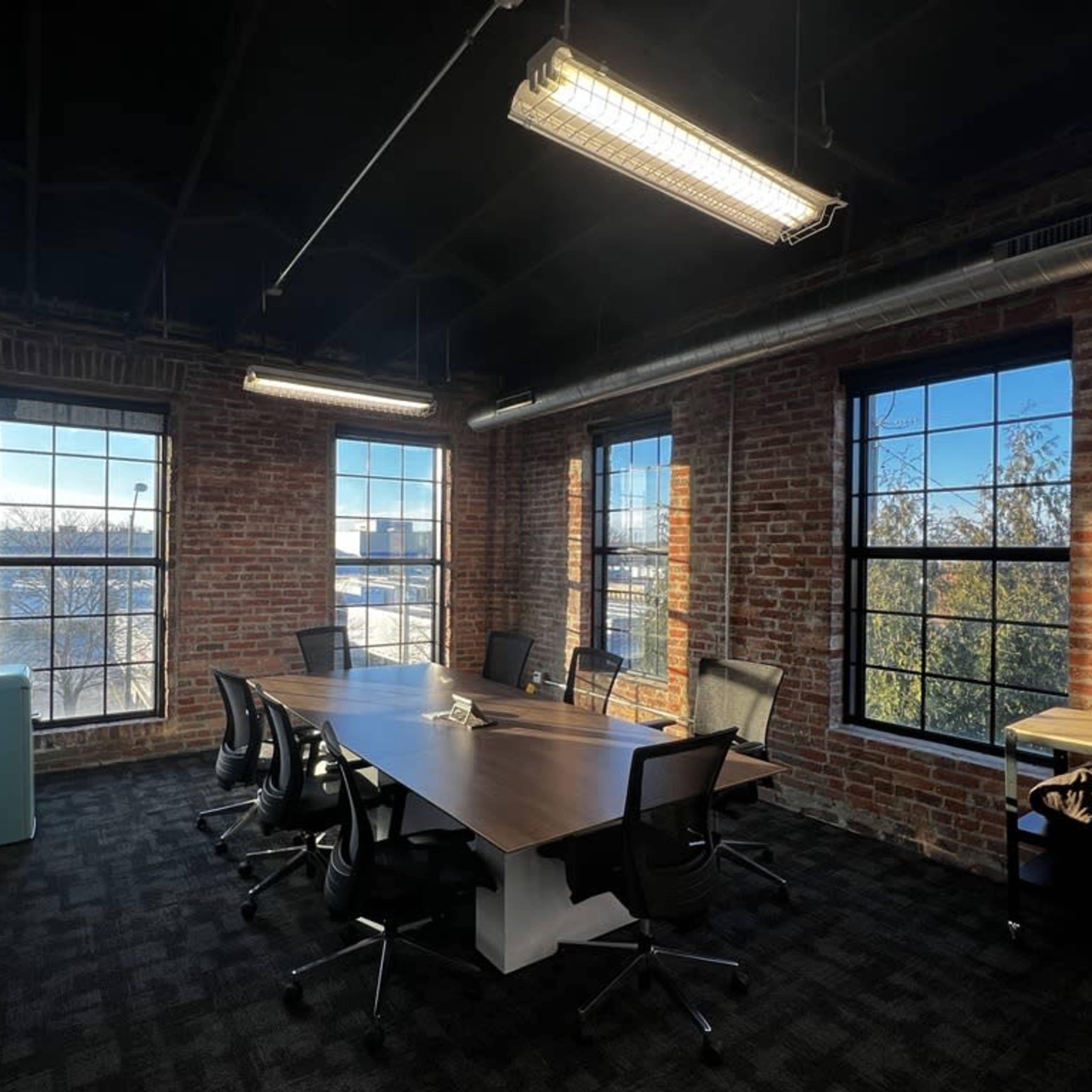 A conference room with a large wooden table surrounded by black chairs, featuring exposed brick walls and large windows that let in natural light.
