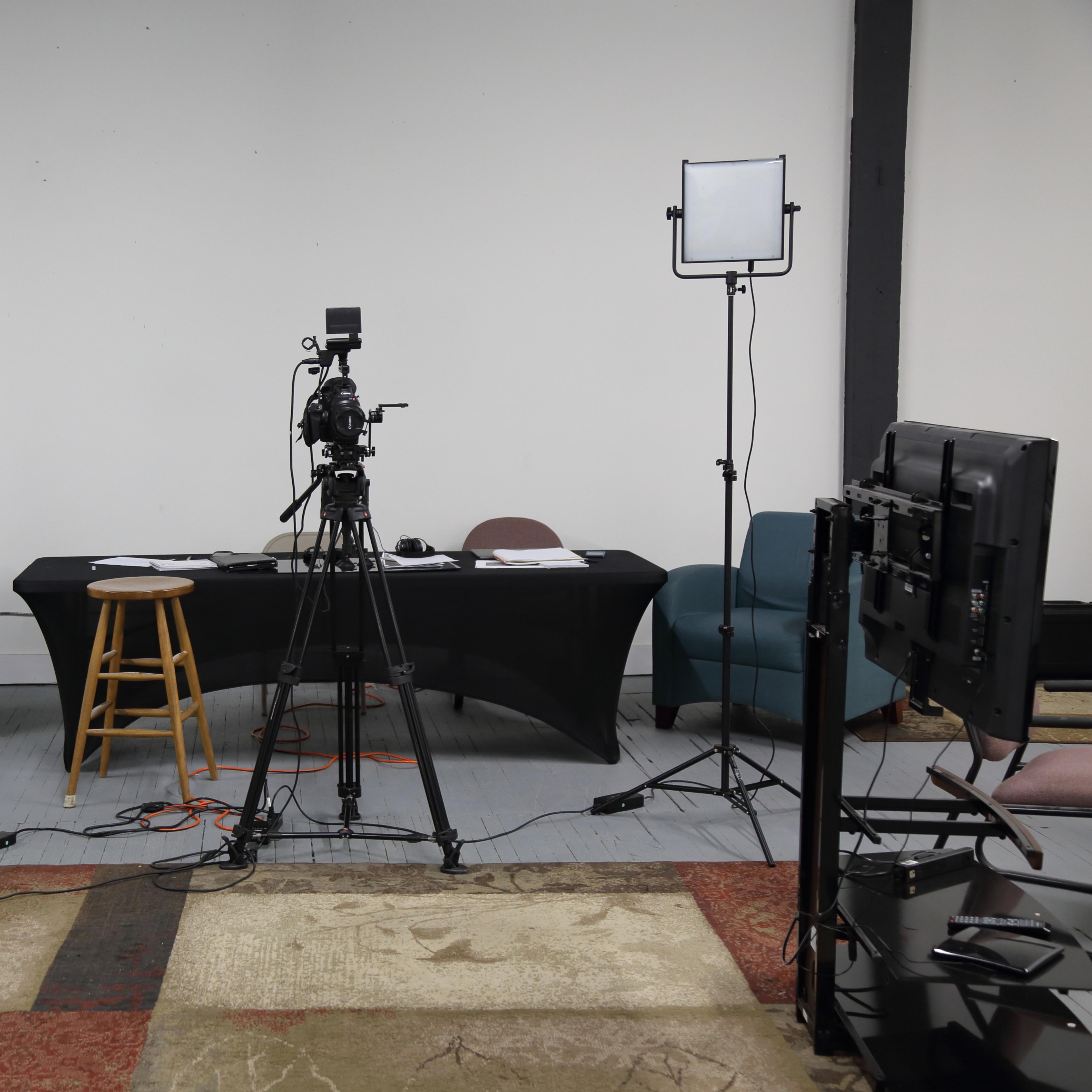 A video recording setup features a table, two chairs, and multiple cameras and lights arranged in a spacious room with a light-colored floor and a patterned rug.