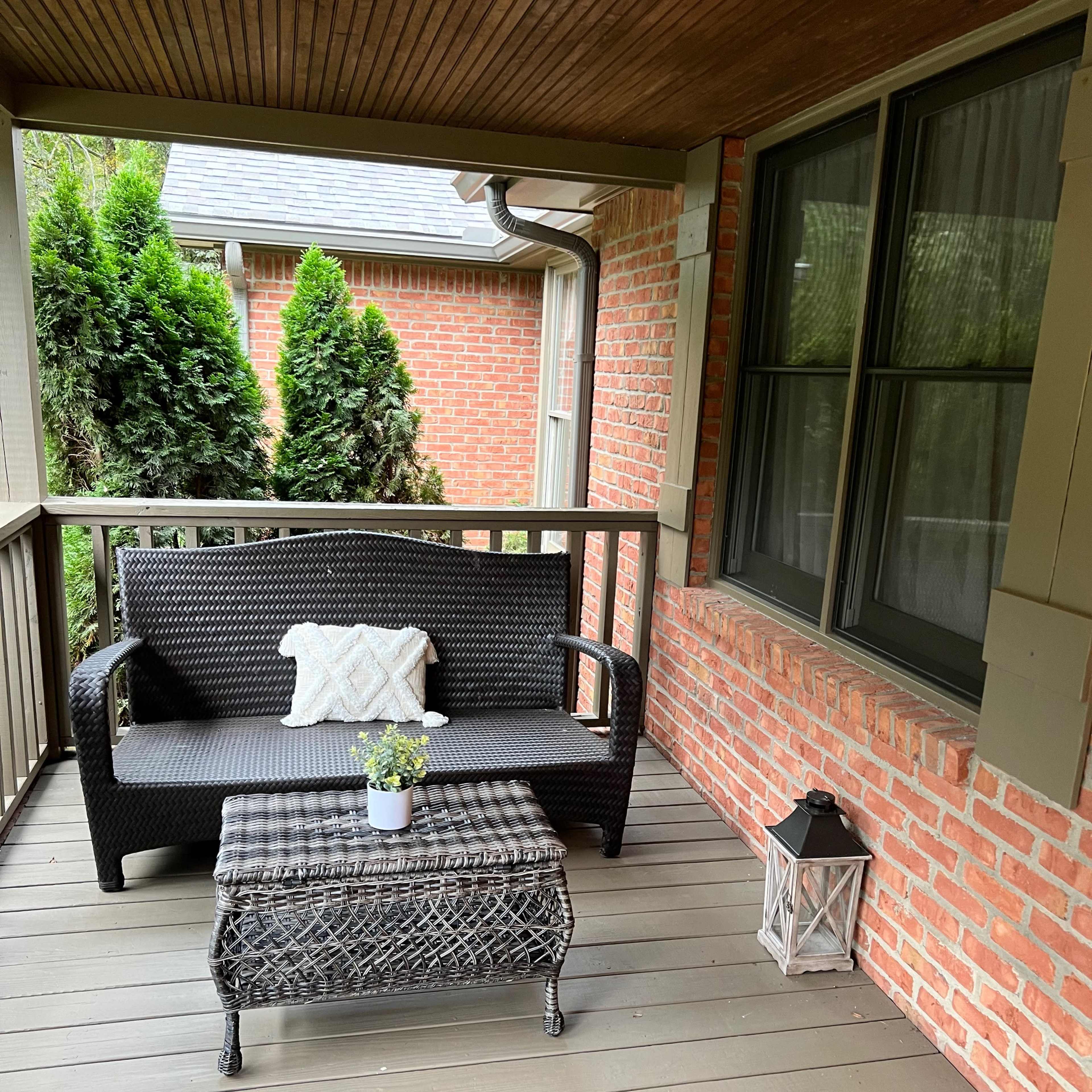 A covered porch with a black wicker sofa, a small table, and a lantern, surrounded by brick walls and greenery.