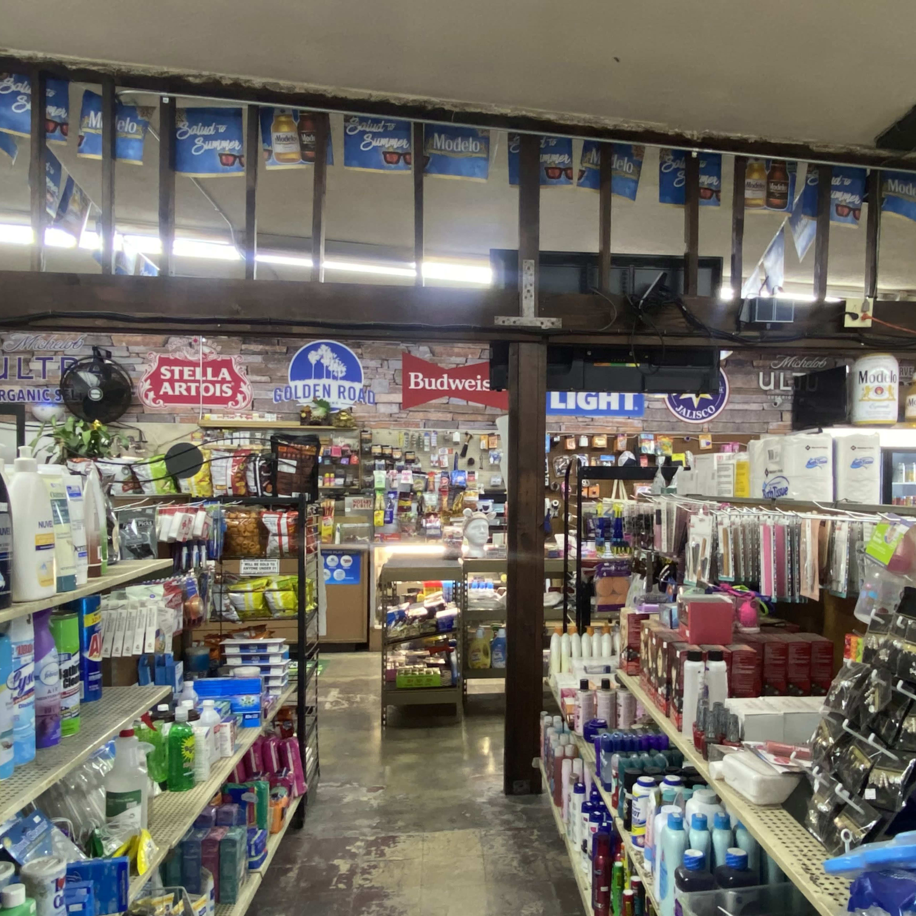 The interior of a convenience store, featuring shelves stocked with various household products and signage for beer brands hanging above.