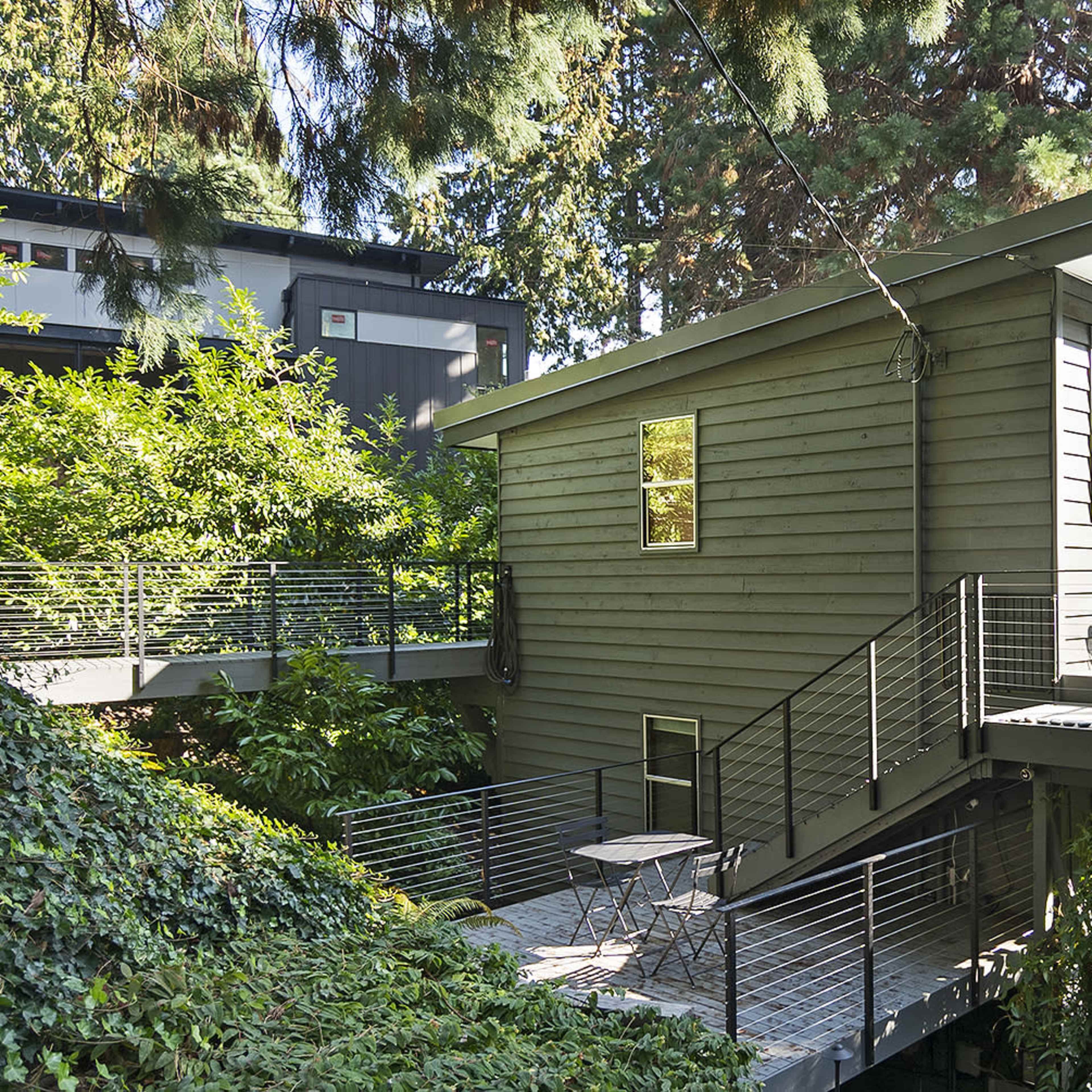 A modern, two-story house surrounded by greenery, featuring a deck with a small table and stairs leading down to the lower level.