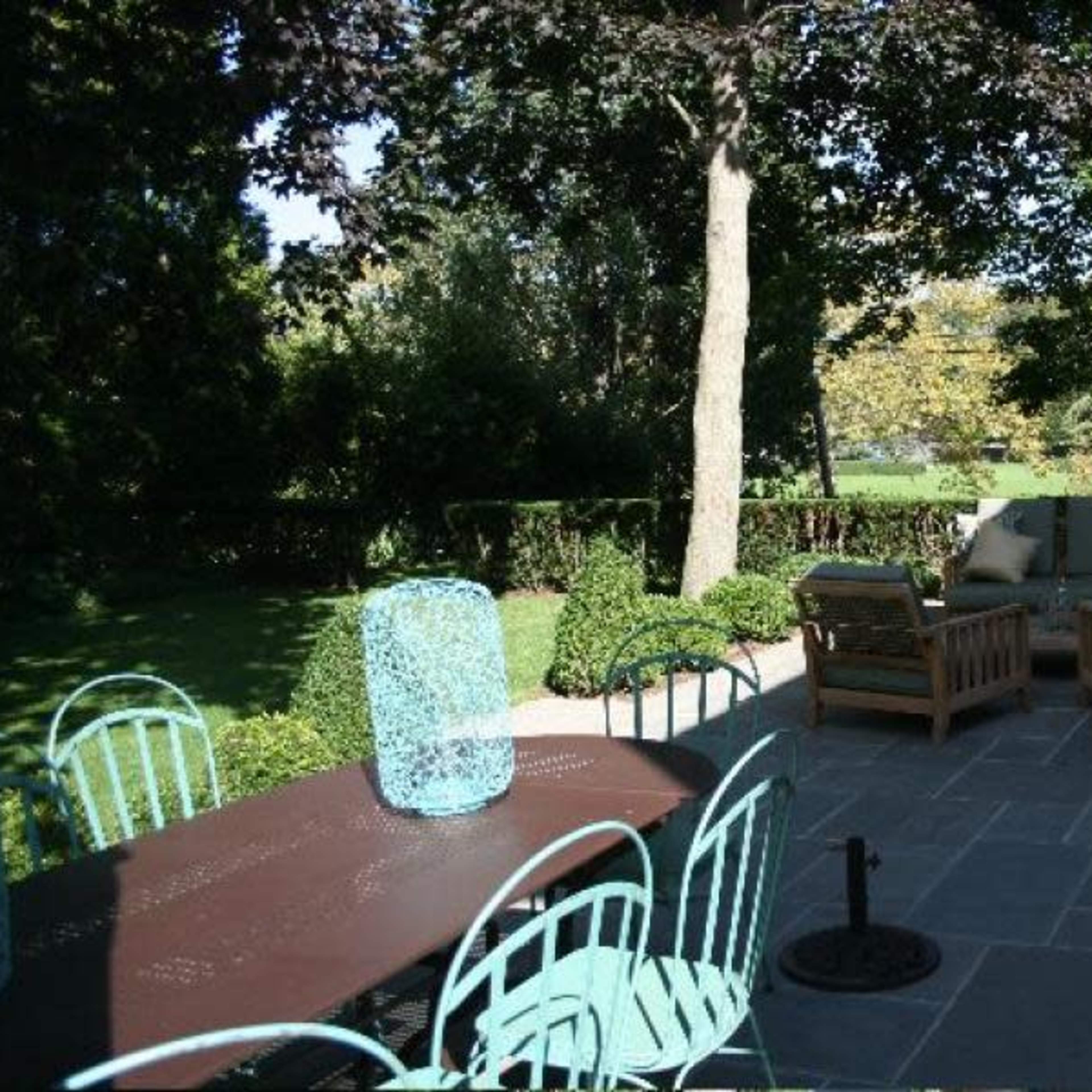 A patio area with a dining table and chairs, along with a seating arrangement featuring wooden chairs and cushions, surrounded by greenery.
