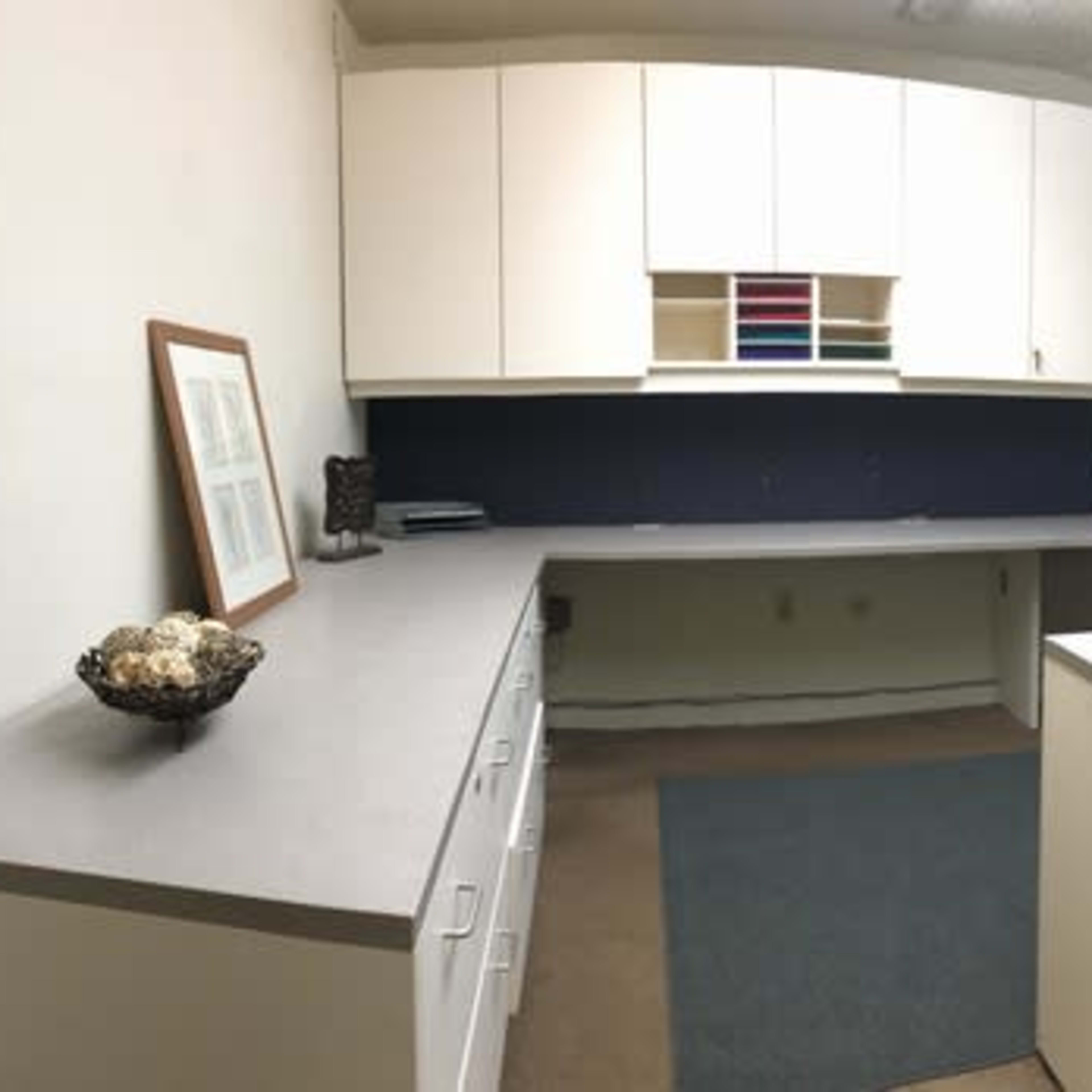 An office space featuring a large L-shaped desk, a black office chair, and a decorative bowl on a table, with shelves above the desk and window blinds.