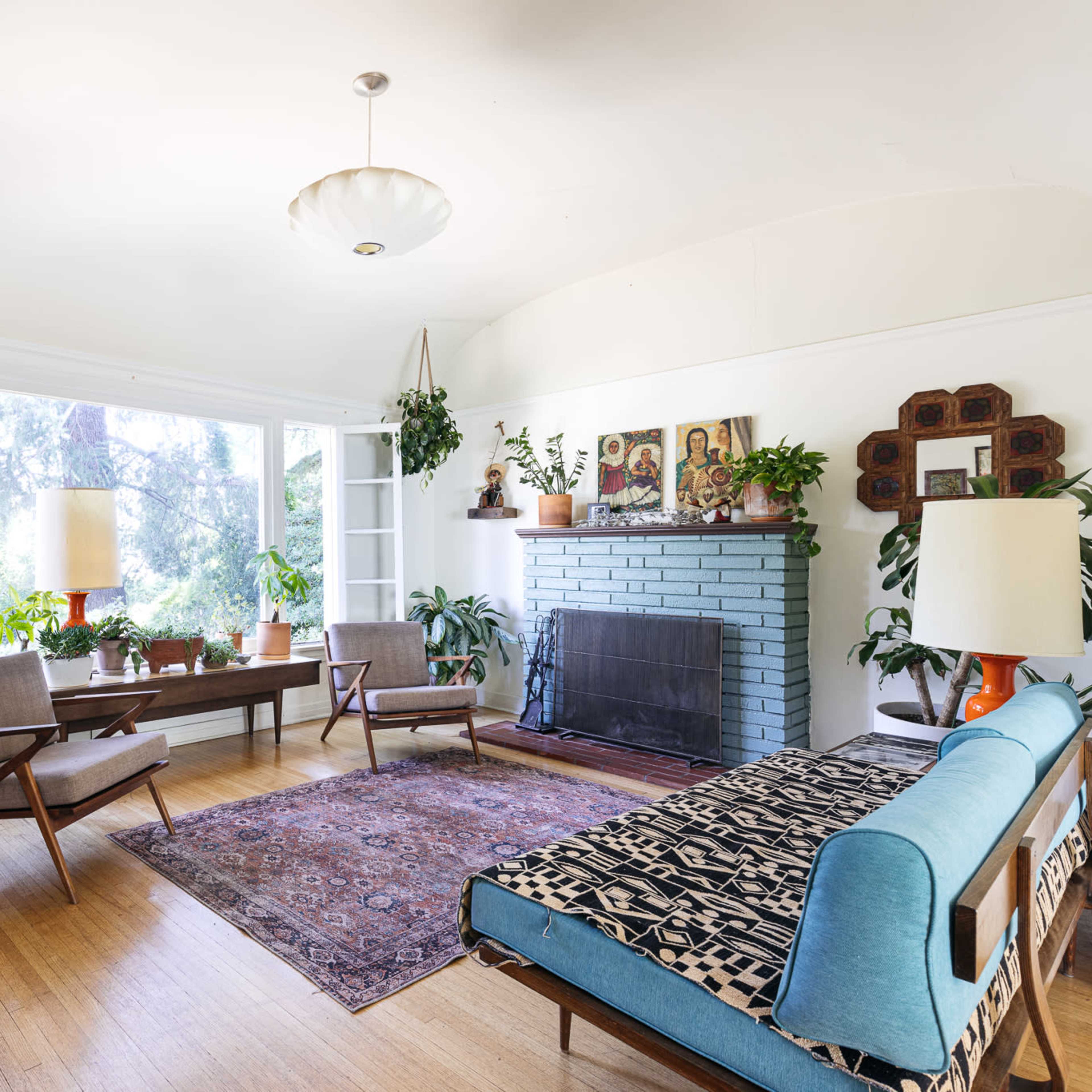 A light-filled living room with a blue sofa, a wooden coffee table, and several potted plants, featuring a fireplace and large windows.
