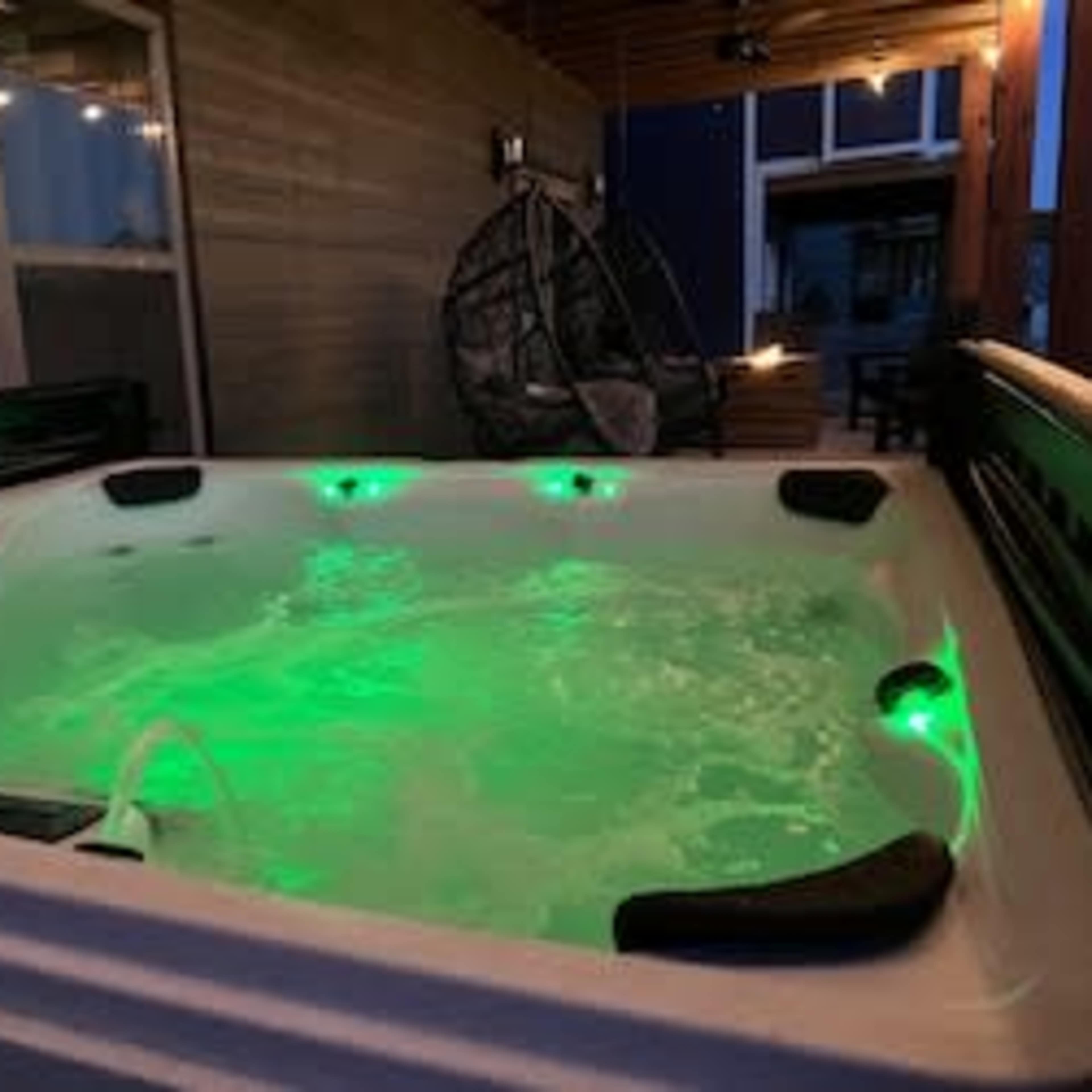 A hot tub with green lighting is situated on a deck with a hanging chair in the background.