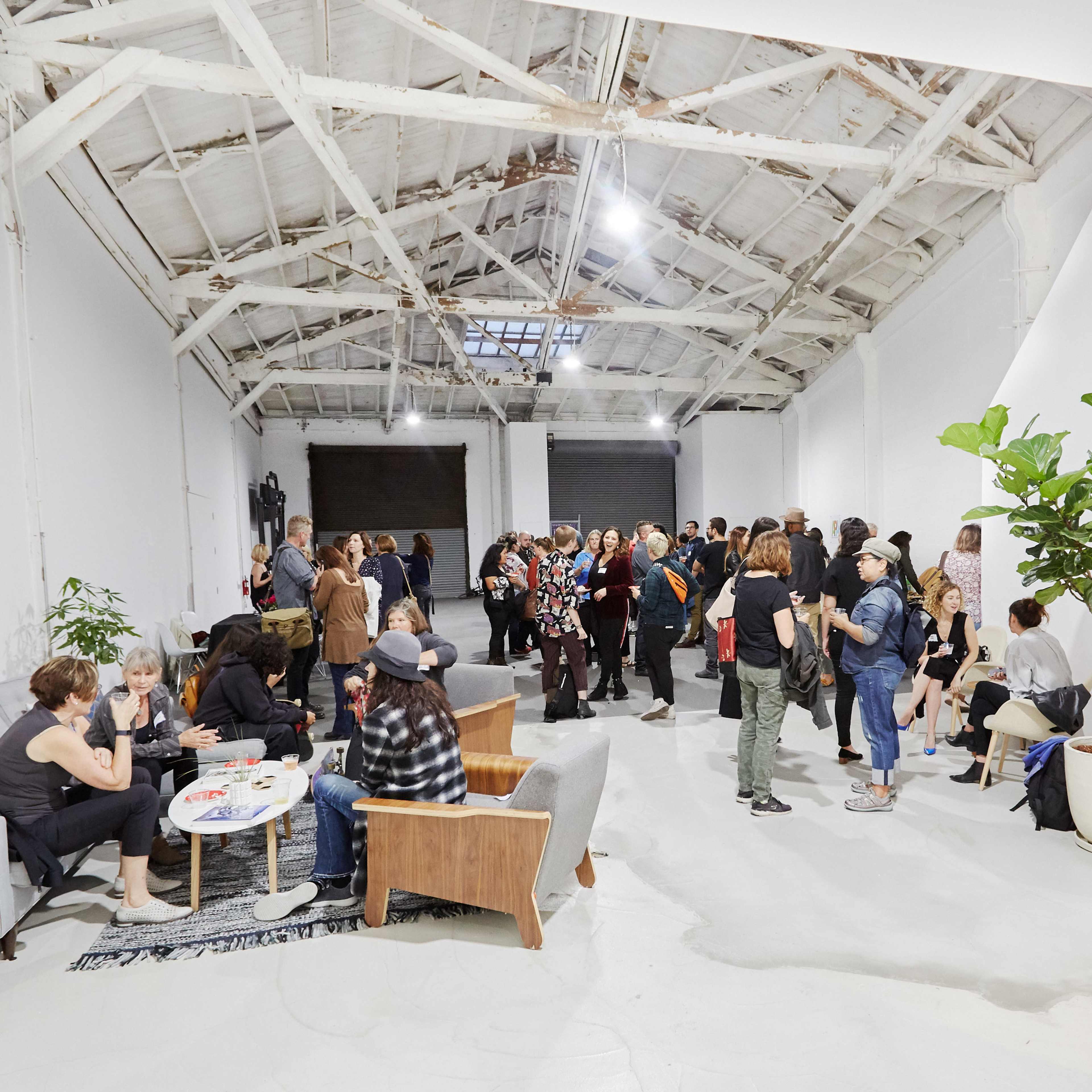 A group of people gather in a spacious, industrial-style room with exposed beams and large windows, socializing and engaging in conversation.