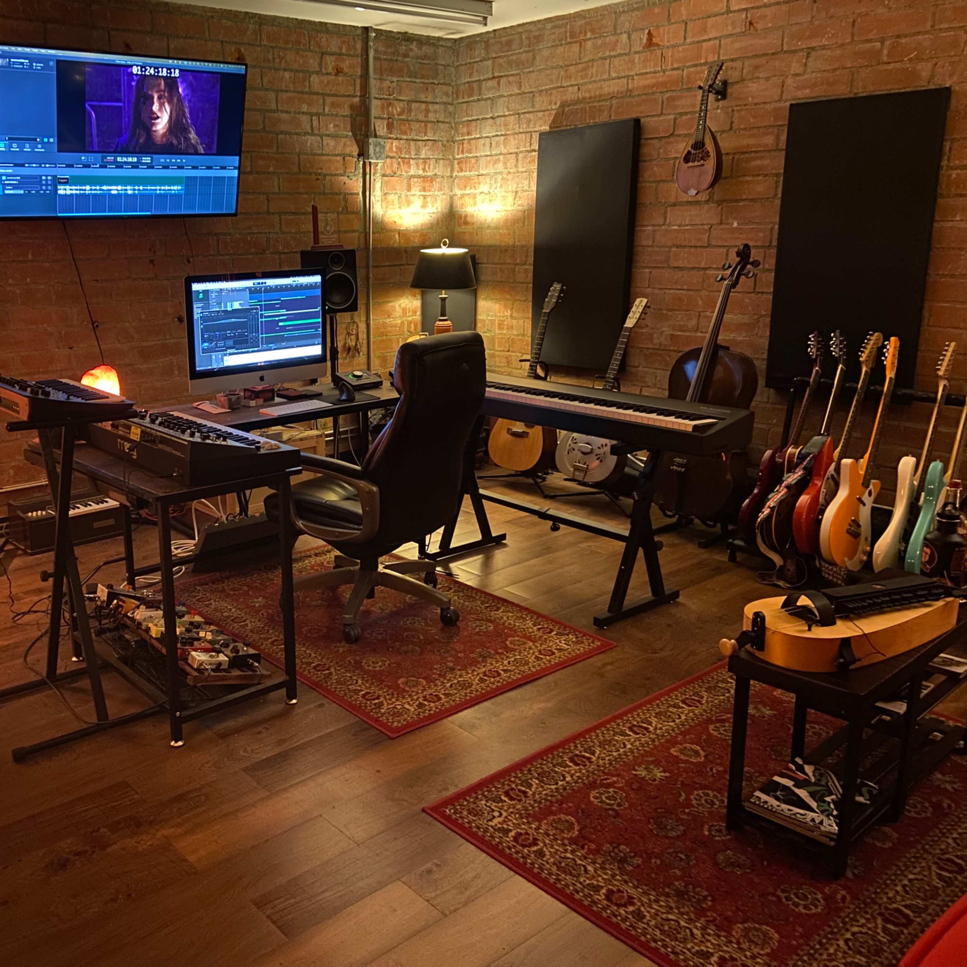 A music studio features a computer desk with monitors, audio equipment, various instruments on display, and exposed brick walls.