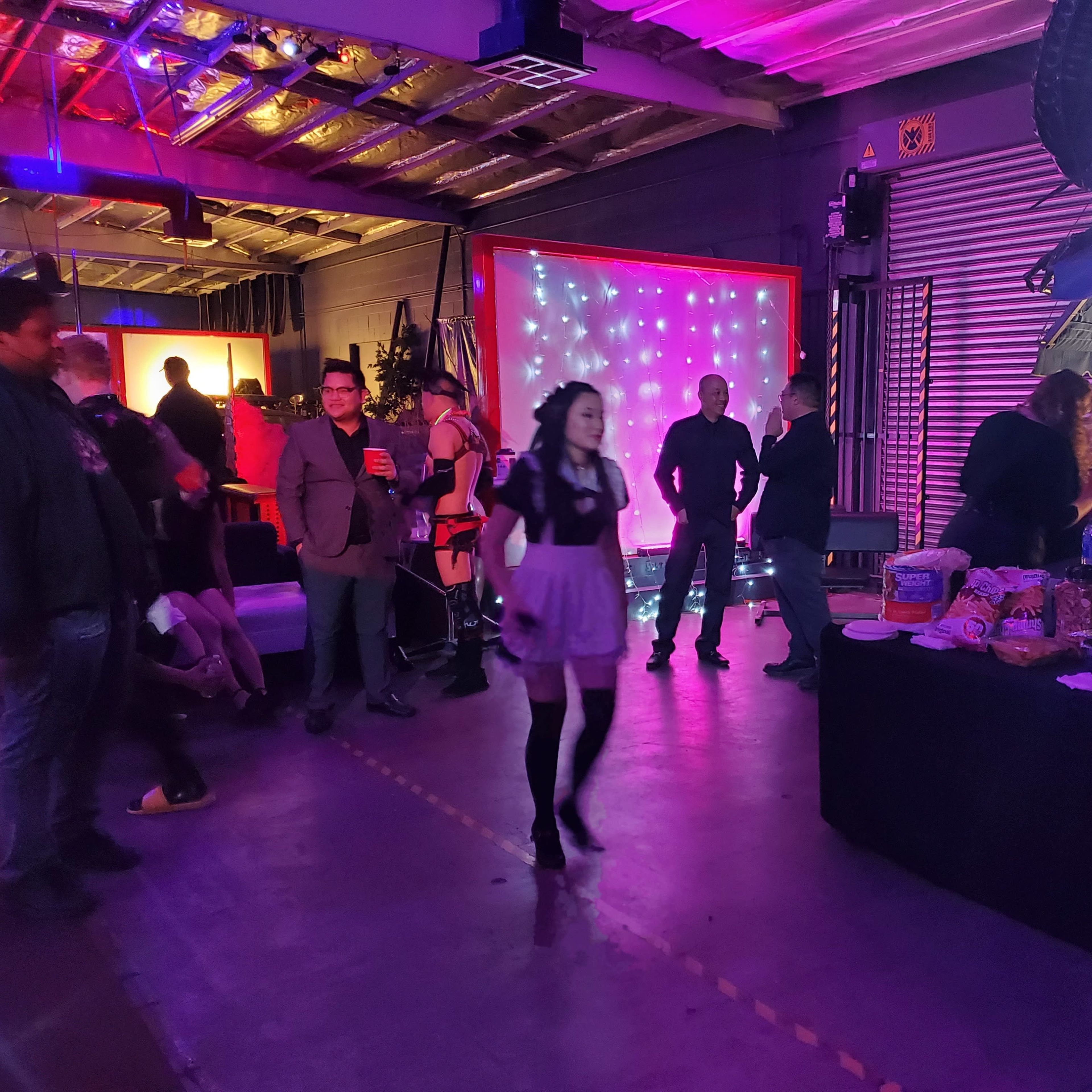 A group of people mingle in a dimly lit venue adorned with colorful lights, while a buffet table displays various food items.