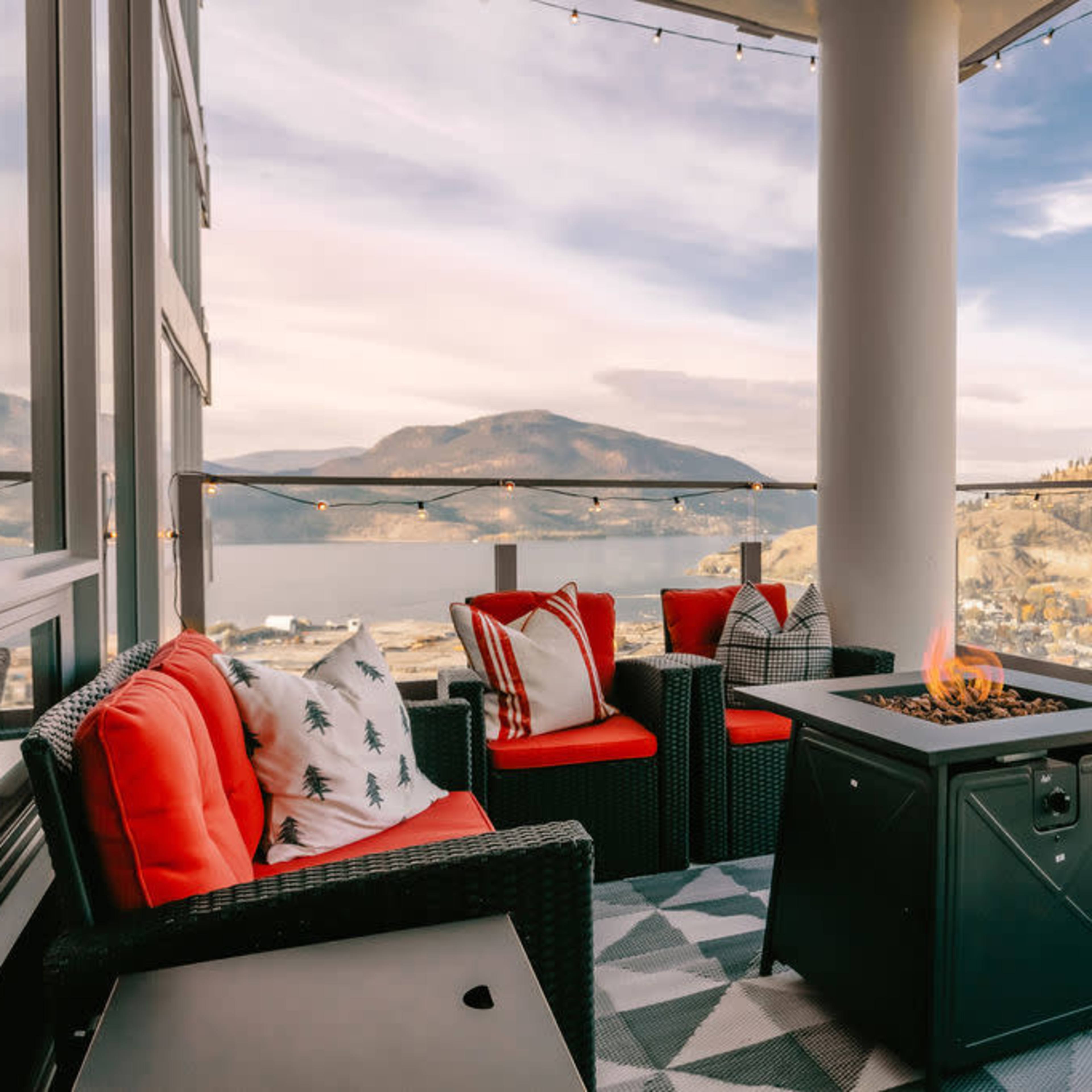 A balcony with two red chairs, a black fire pit, and a view of mountains and a lake in the background.