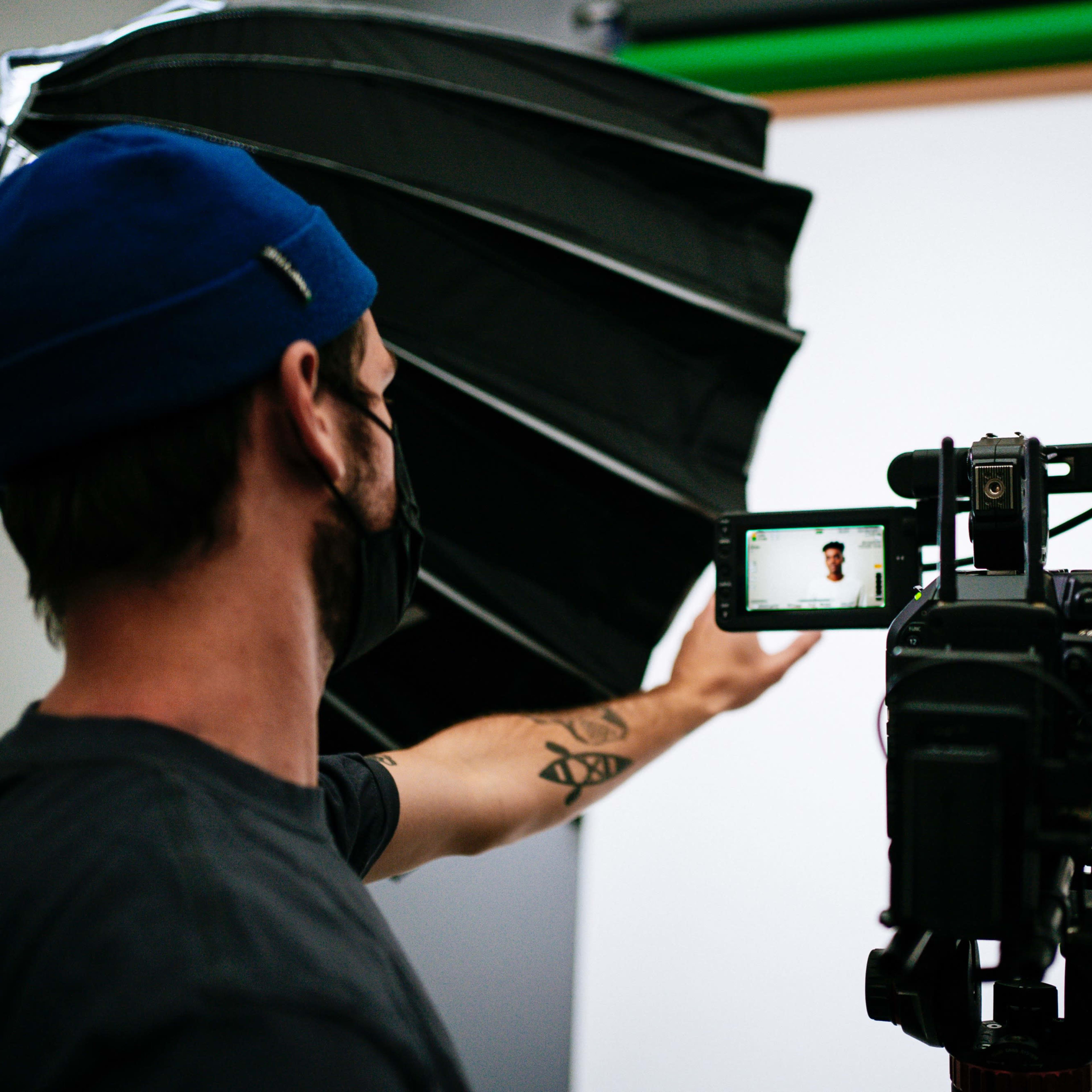 A photographer in a blue cap and mask holds a camera directed at a smiling subject in a white shirt against a plain backdrop.