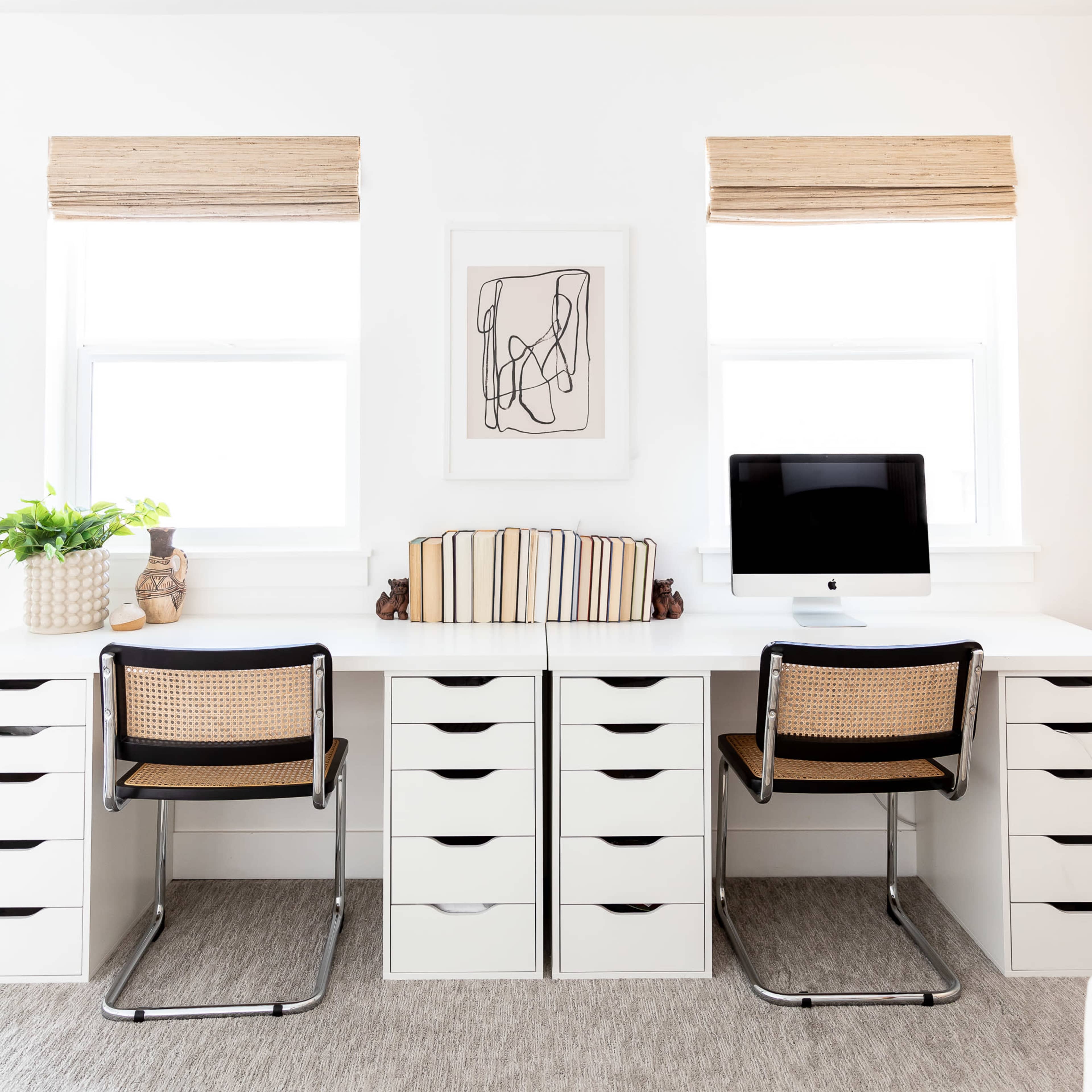 A modern home office features two desks with matching chairs, surrounded by storage drawers and illuminated by natural light from two windows with shades.