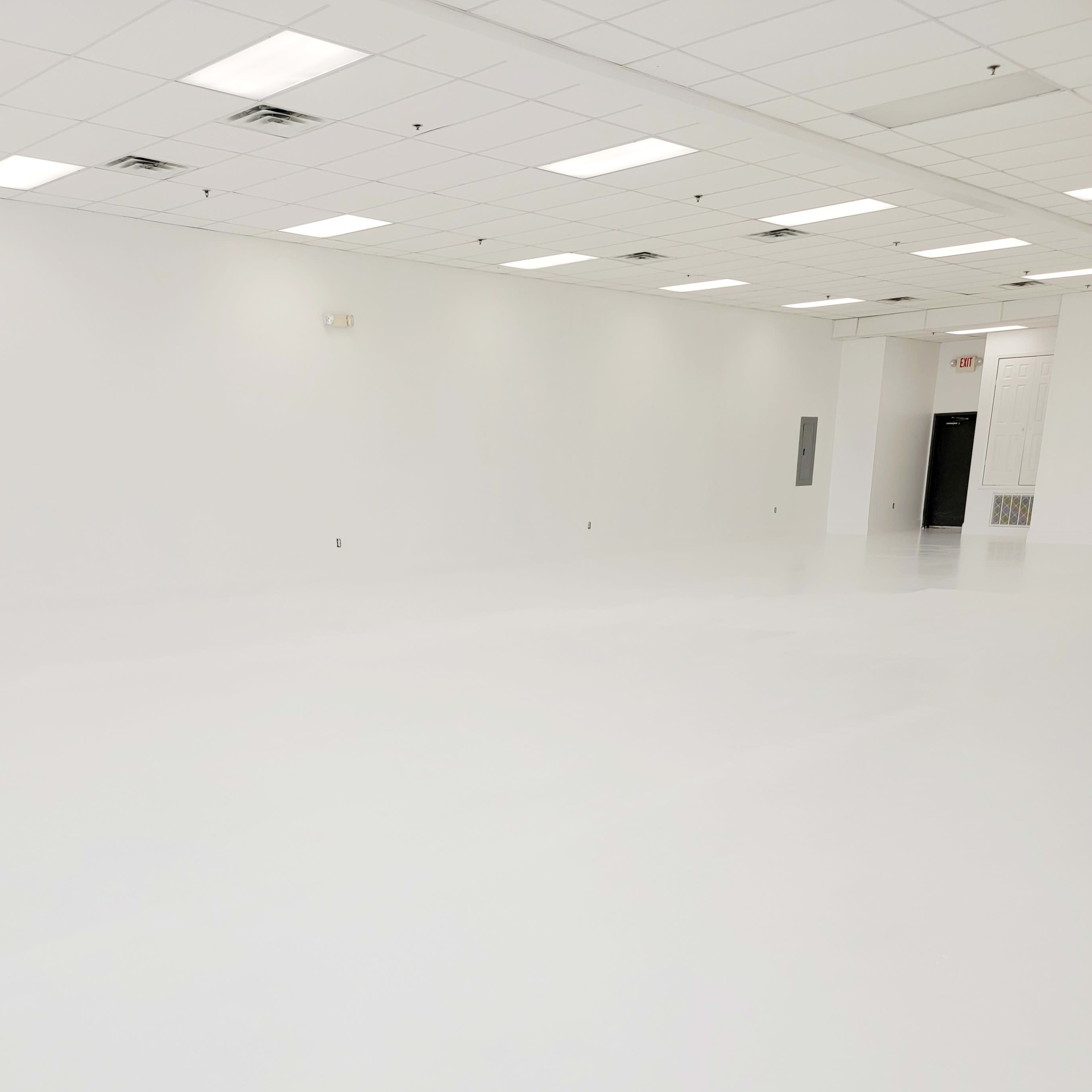 An empty room with white walls and a white floor, featuring a few ceiling lights and a single door on one side.
