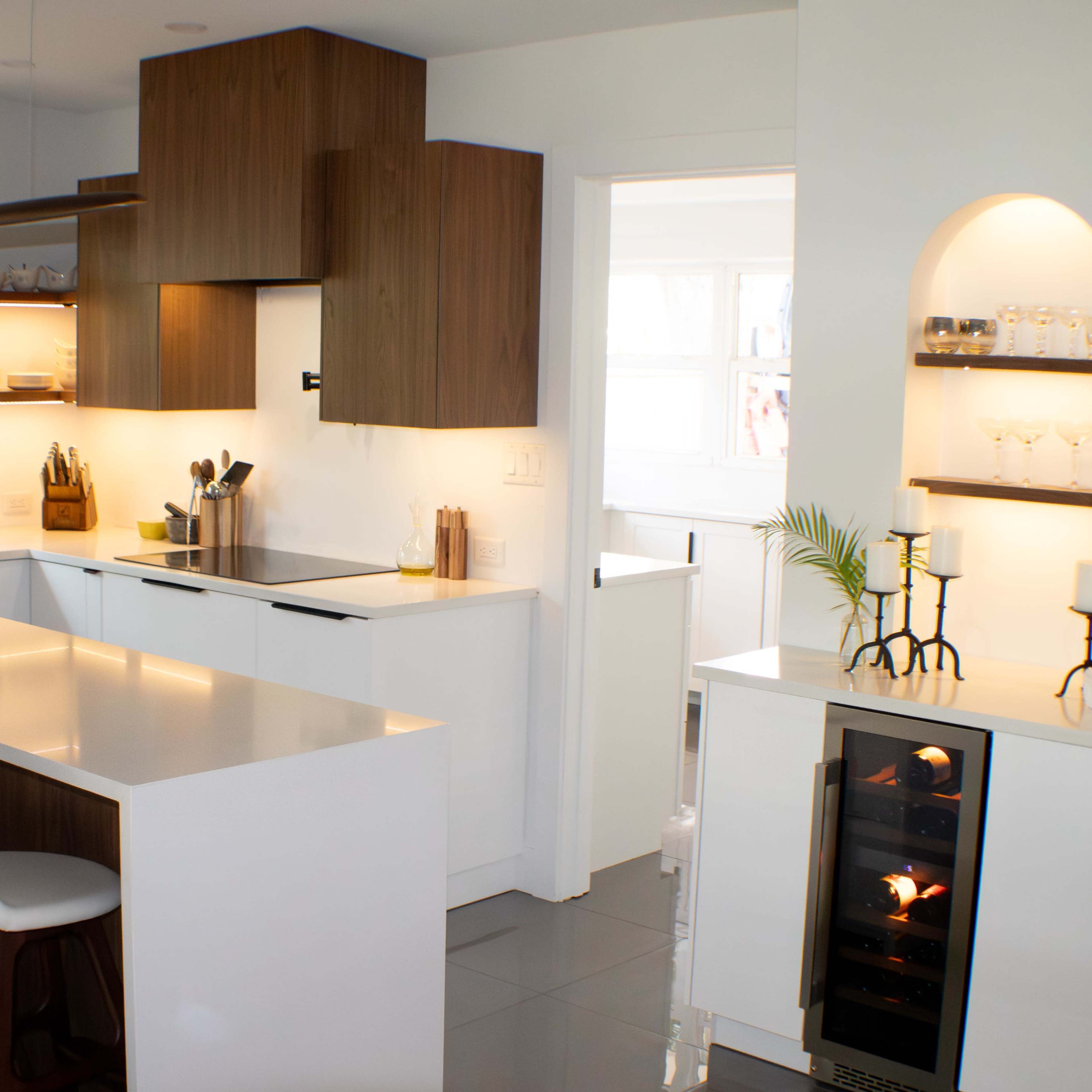 A modern kitchen features a central island with three stools, sleek cabinetry, and a wine cooler beside an arched nook with glassware.