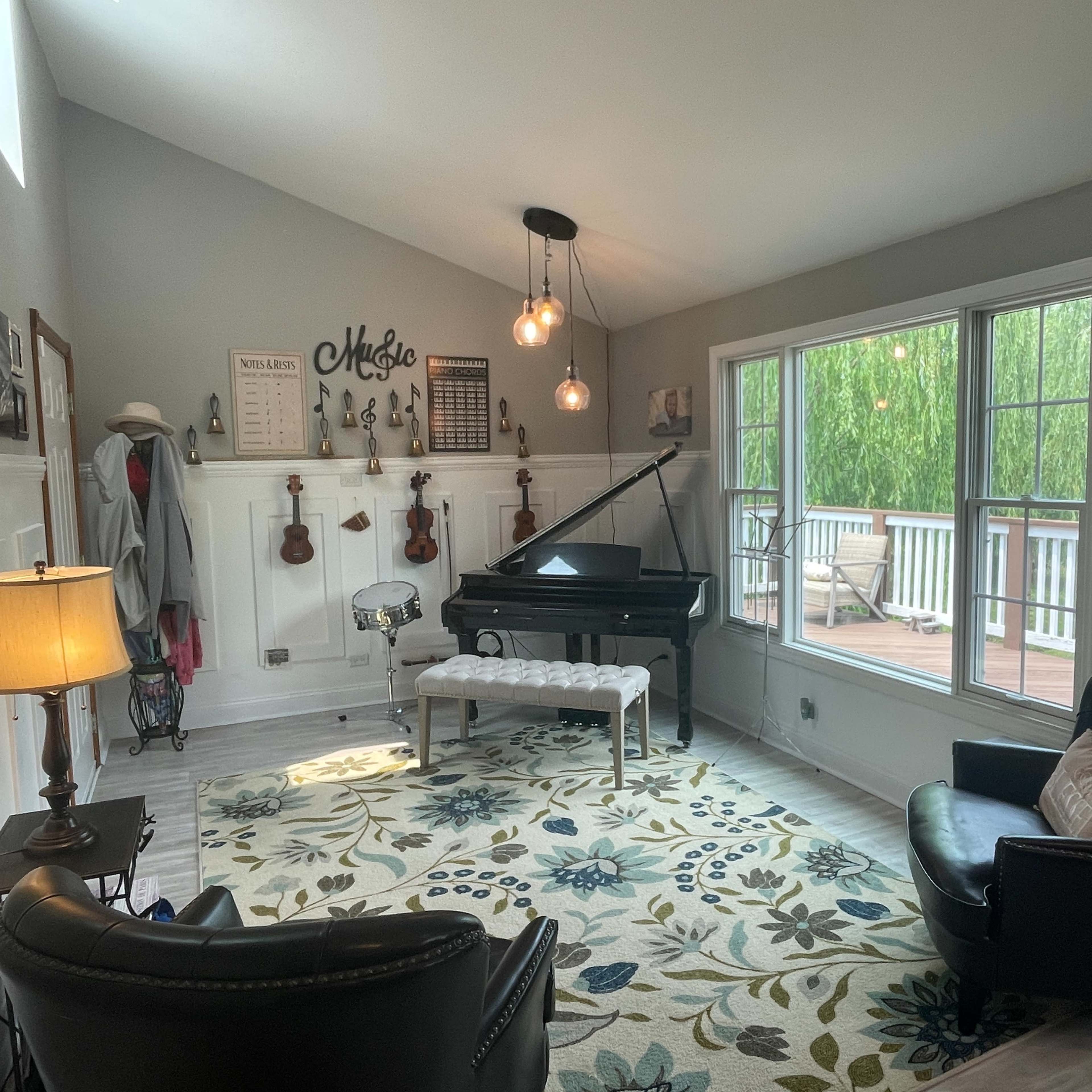 The room features a black grand piano, two black chairs, and a patterned rug, with large windows overlooking a deck and greenery.