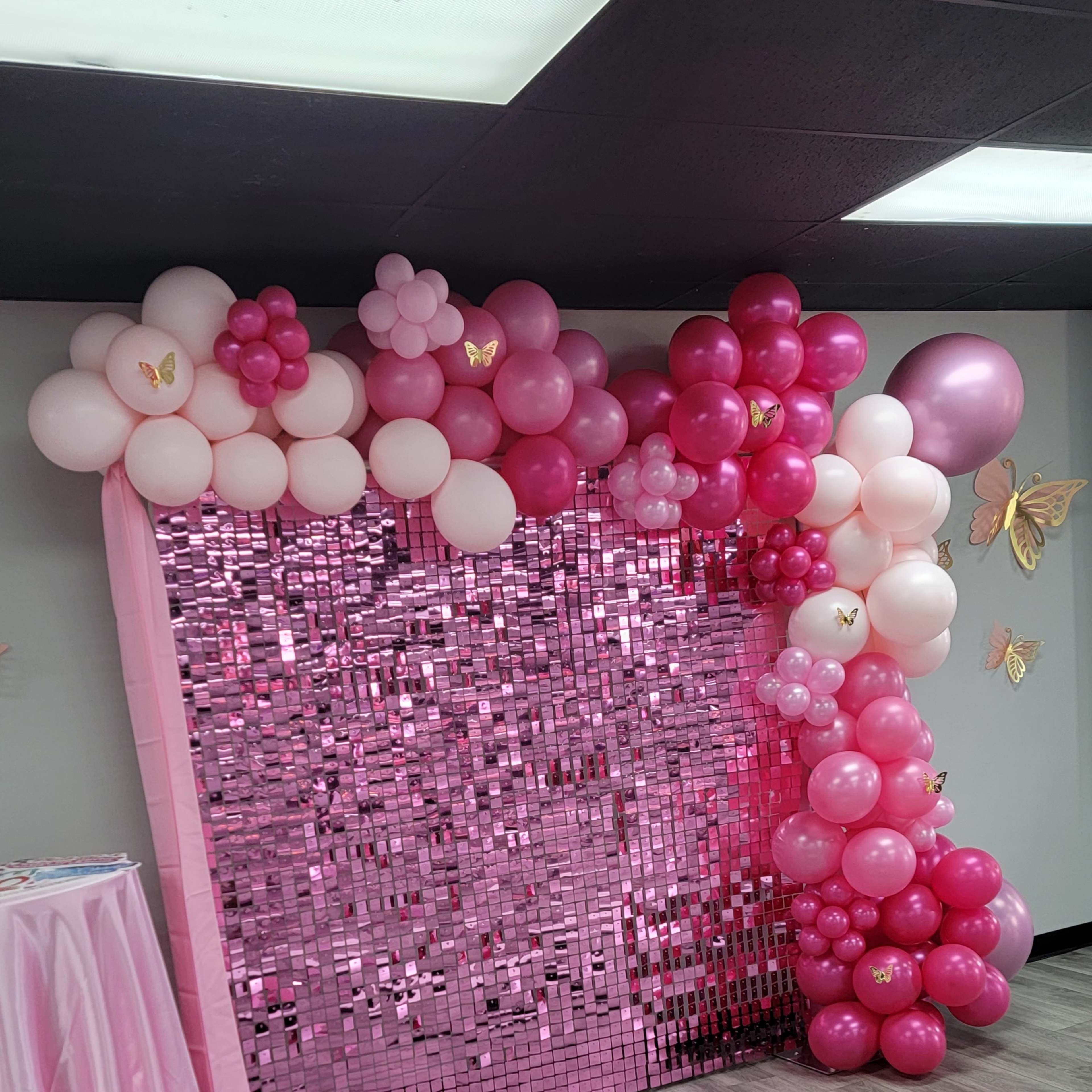 The scene features a backdrop made of pink balloons and shiny decorations, alongside a pink tablecloth draped over a table.