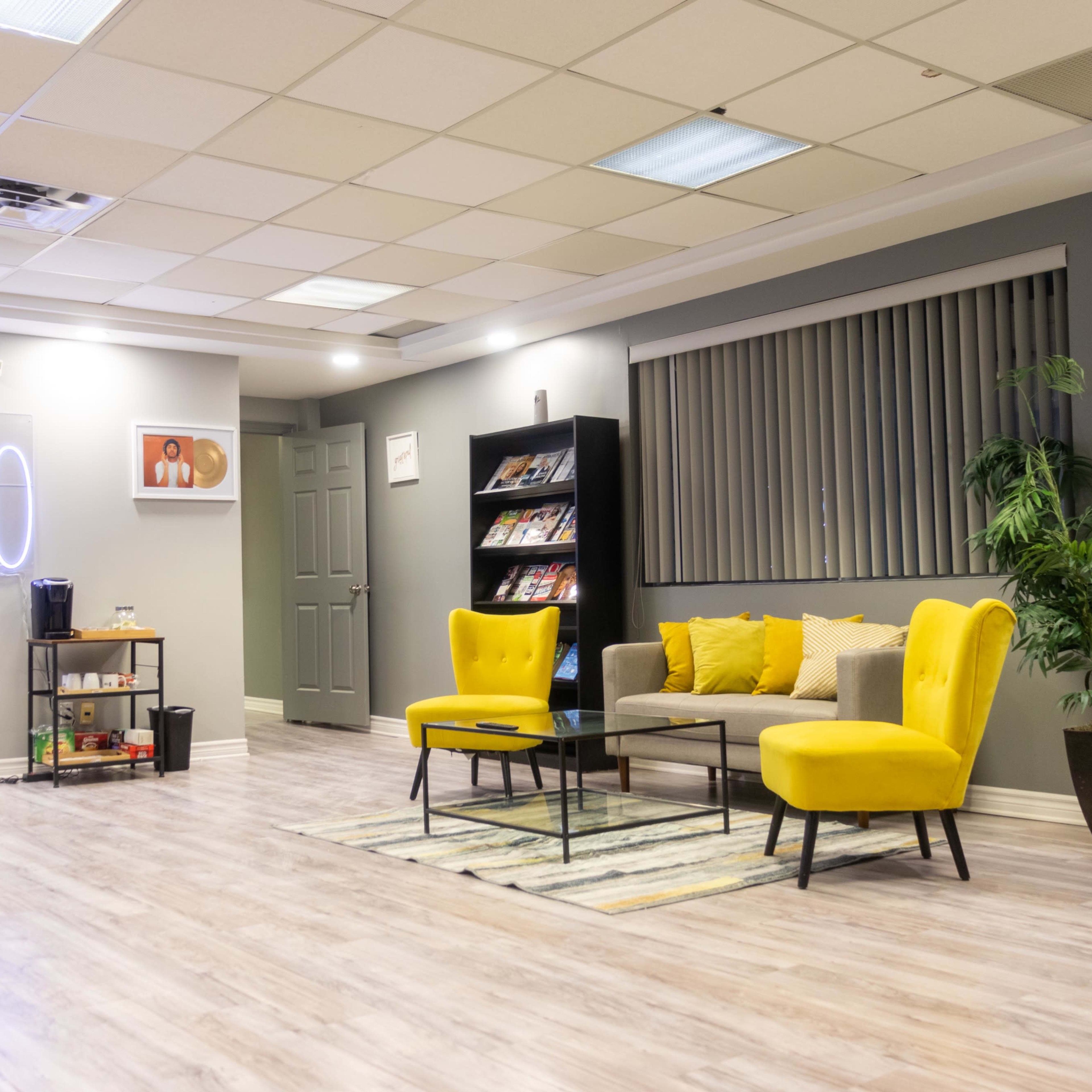 A modern studio space with neon signage reading "ESTUDIO," two yellow chairs, a gray sofa, and a bookshelf filled with magazines.