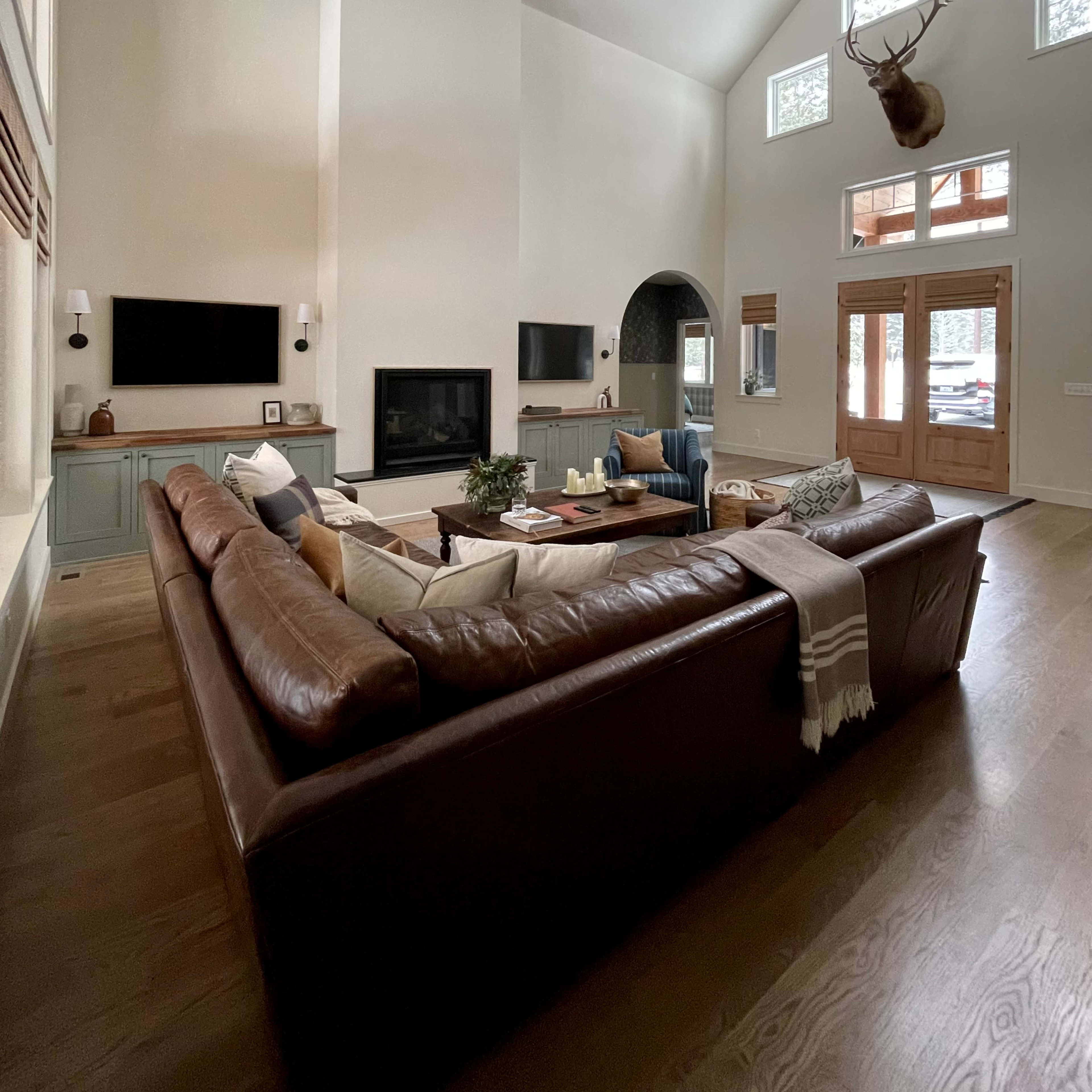 A spacious living room features a large leather sectional sofa, two wall-mounted televisions, and tall windows that fill the space with light.