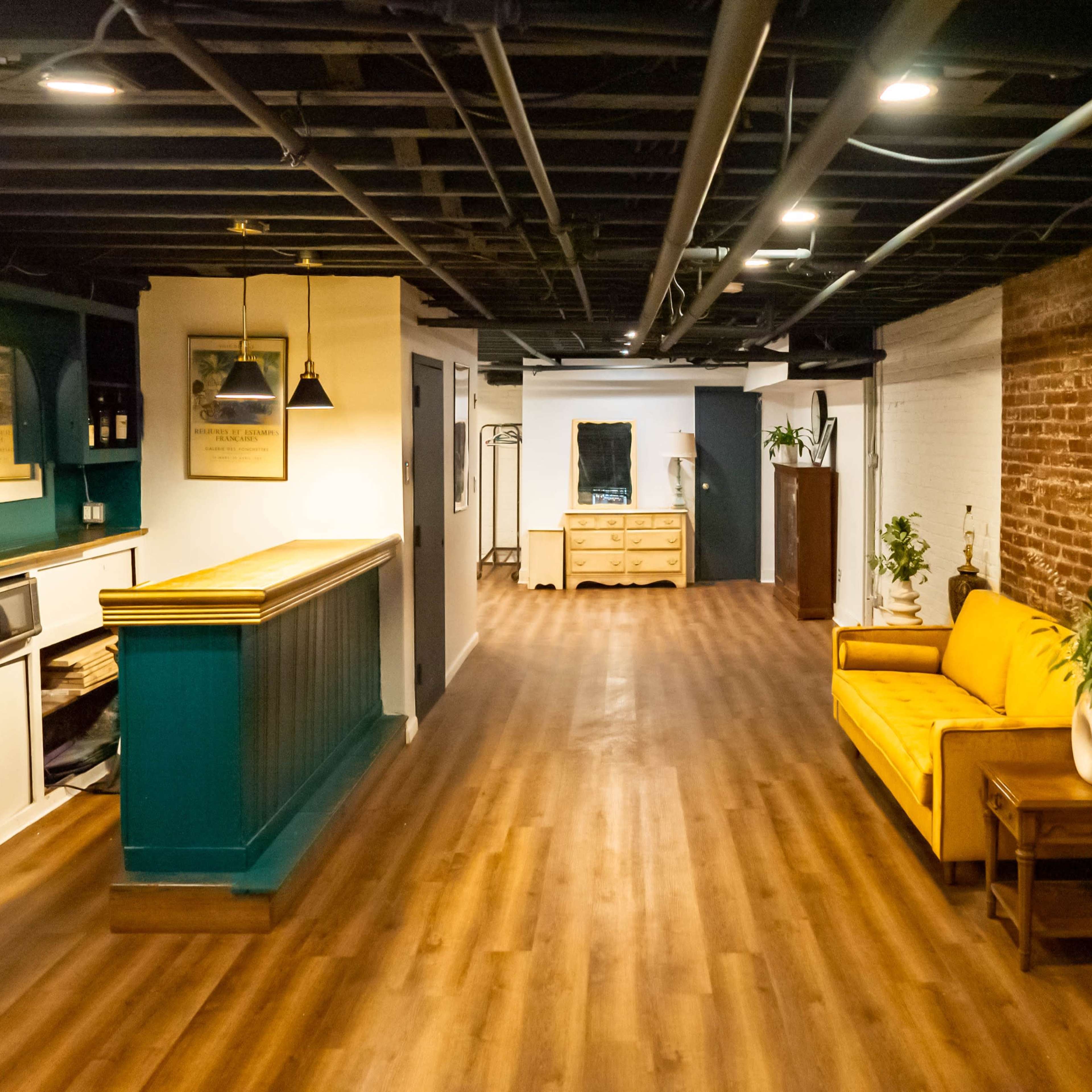 A modern interior space featuring a long, narrow hallway with wooden flooring, a yellow sofa, exposed brick walls, and a teal accent wall with shelves.