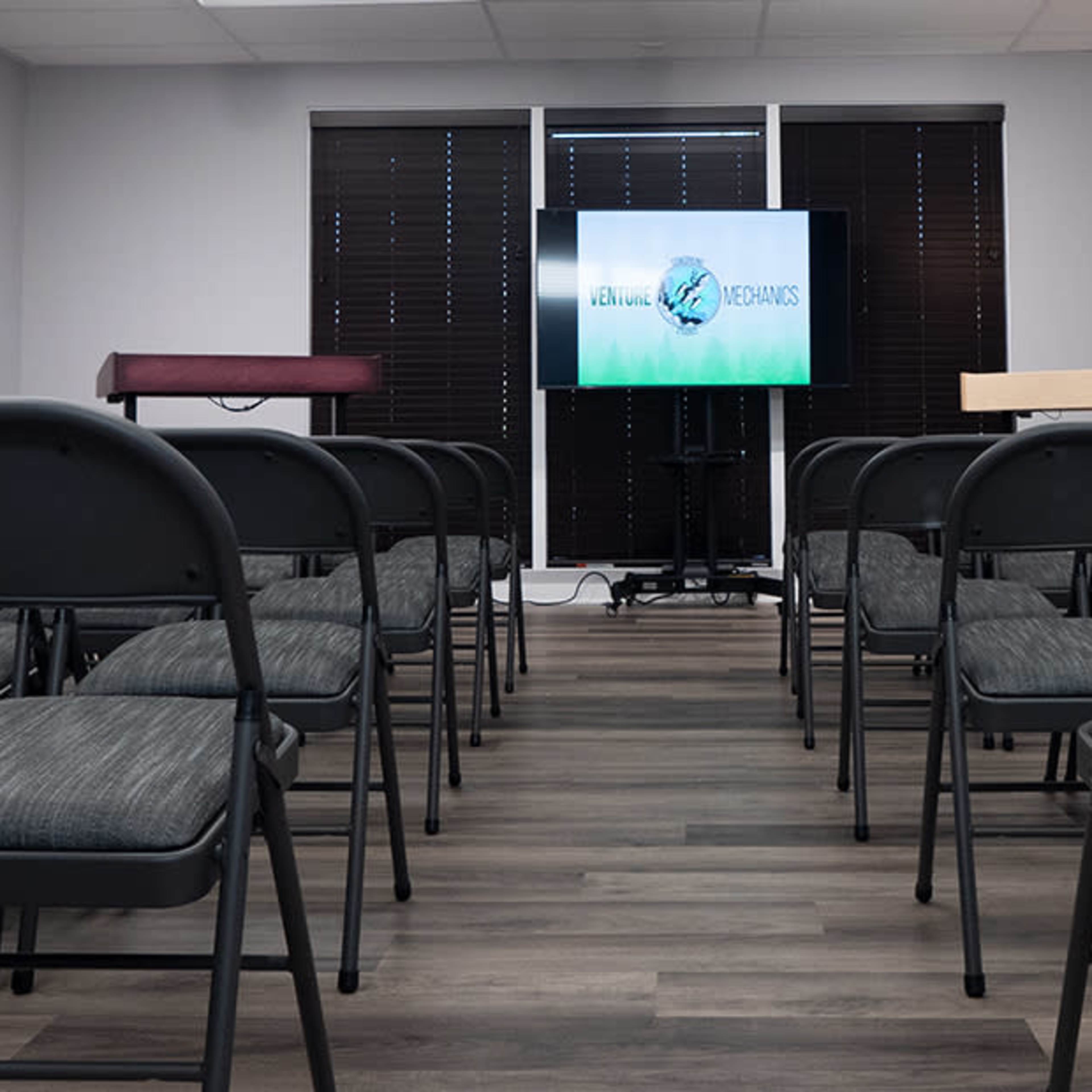 A meeting room with rows of folding chairs facing a screen displaying the logo of a company or organization.