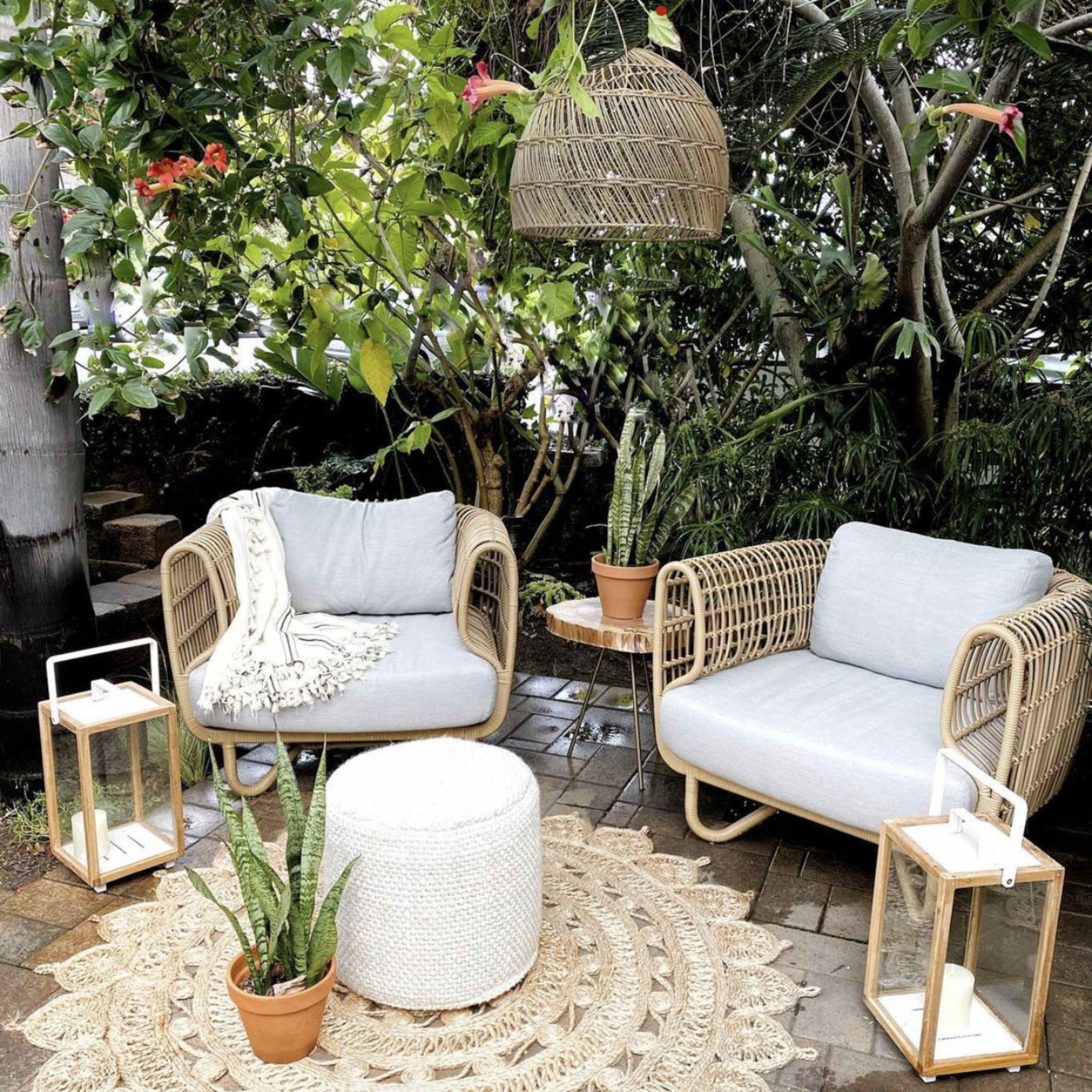 A cozy outdoor seating area features two rattan chairs with gray cushions, a round white pouf, and decorative lanterns arranged on a patterned rug surrounded by greenery.