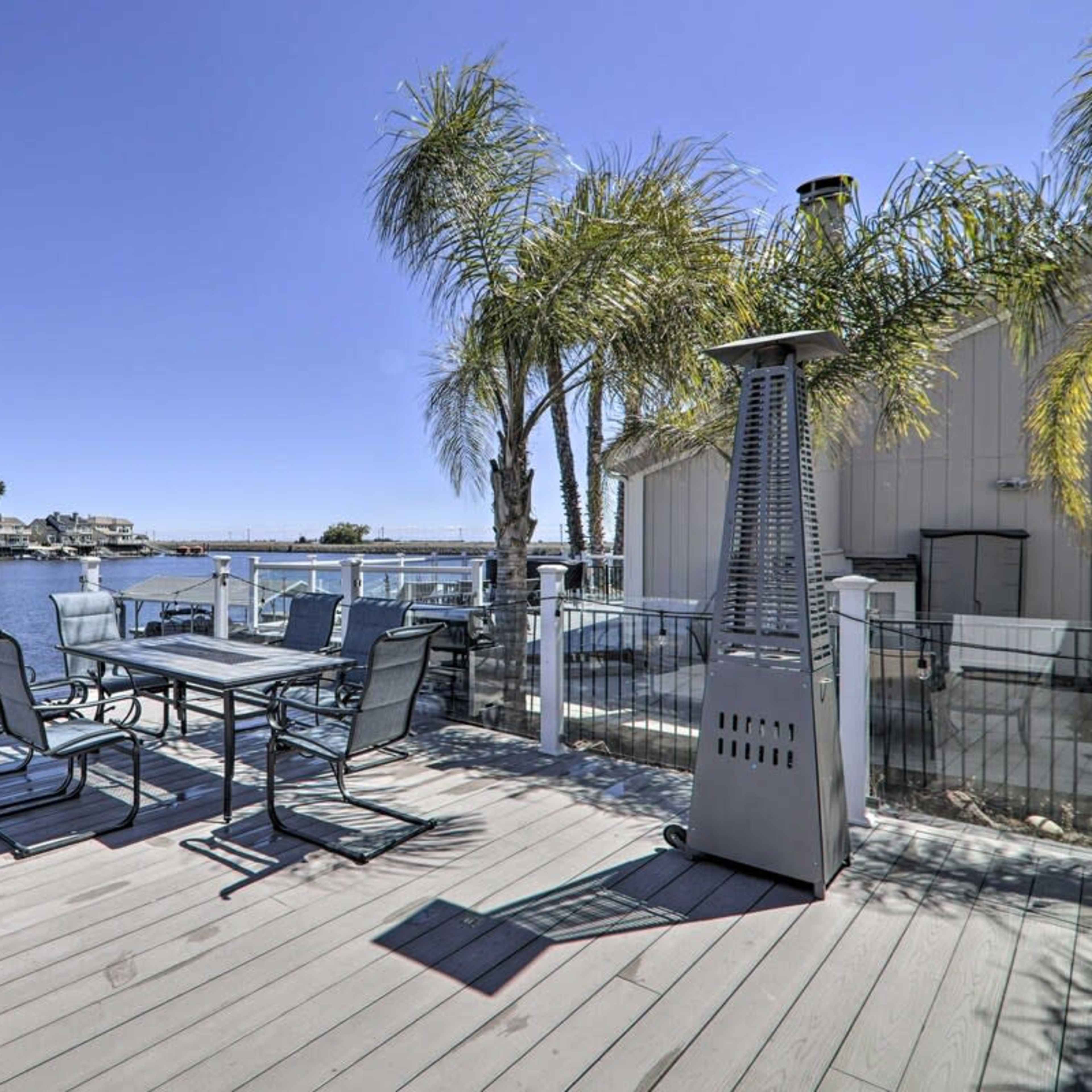 A wooden deck overlooks a calm waterfront with palm trees and dining furniture.