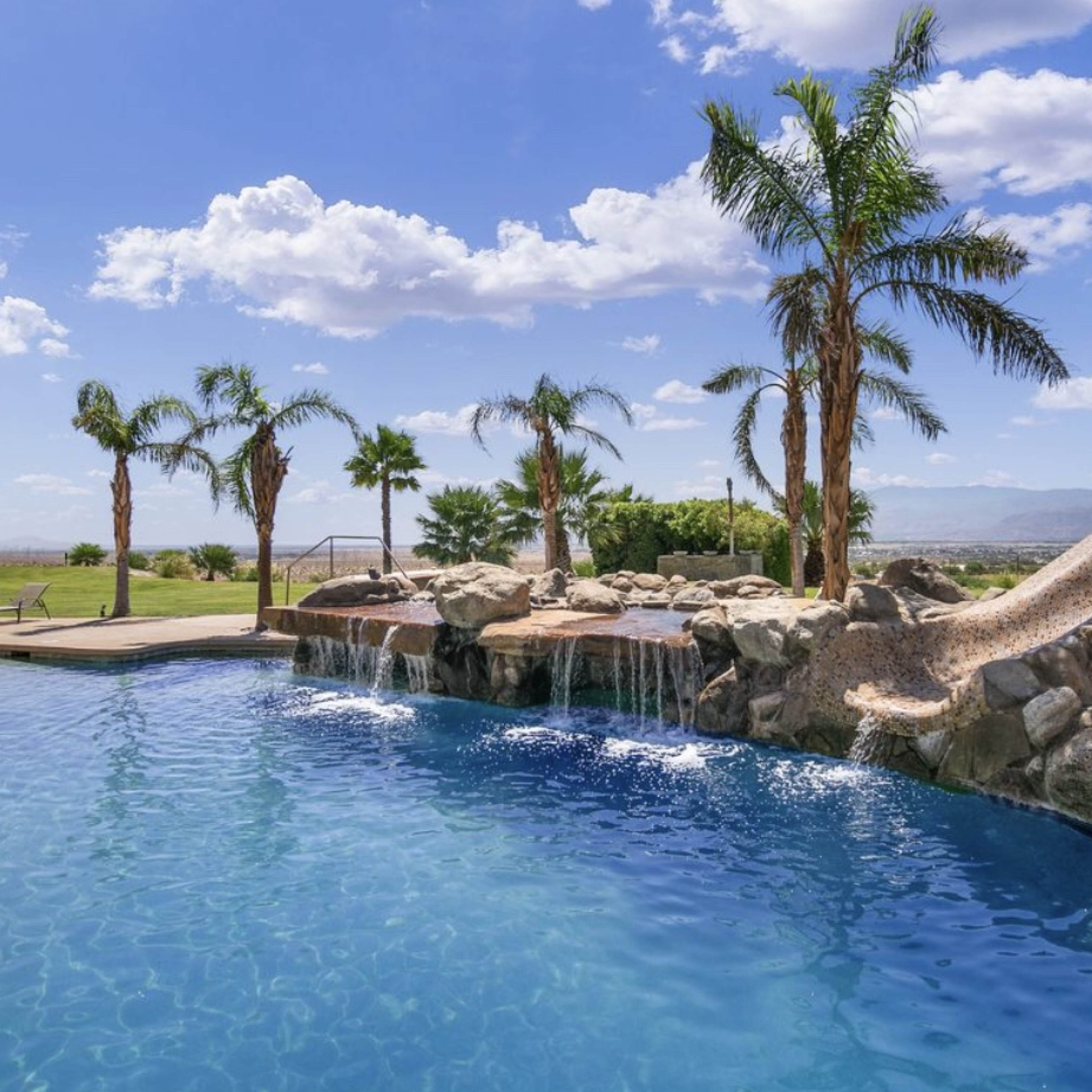 A swimming pool with a waterslide and palm trees, set against a backdrop of blue skies and distant mountains.