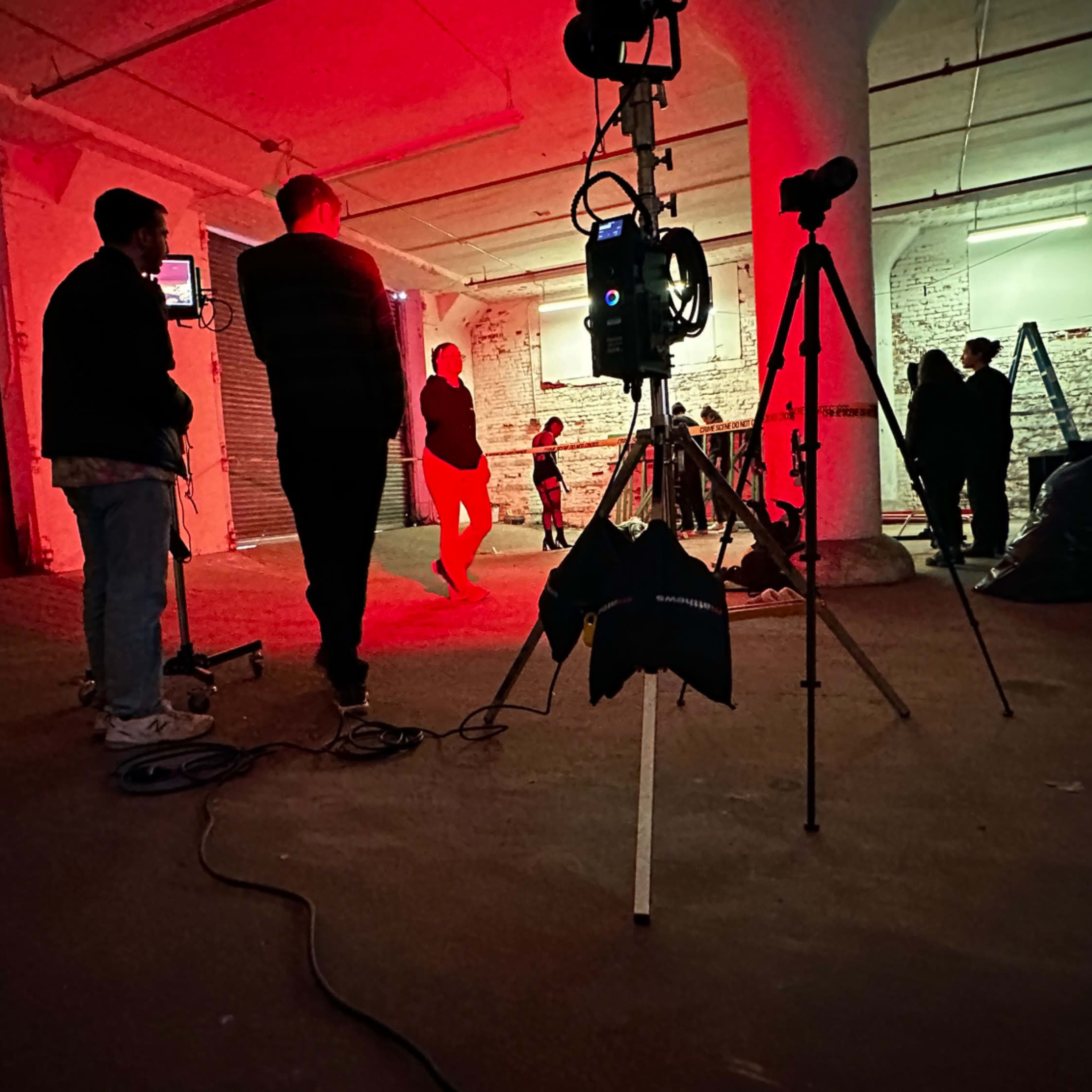 A film crew sets up equipment in a dimly lit warehouse while performers rehearse under red lighting.
