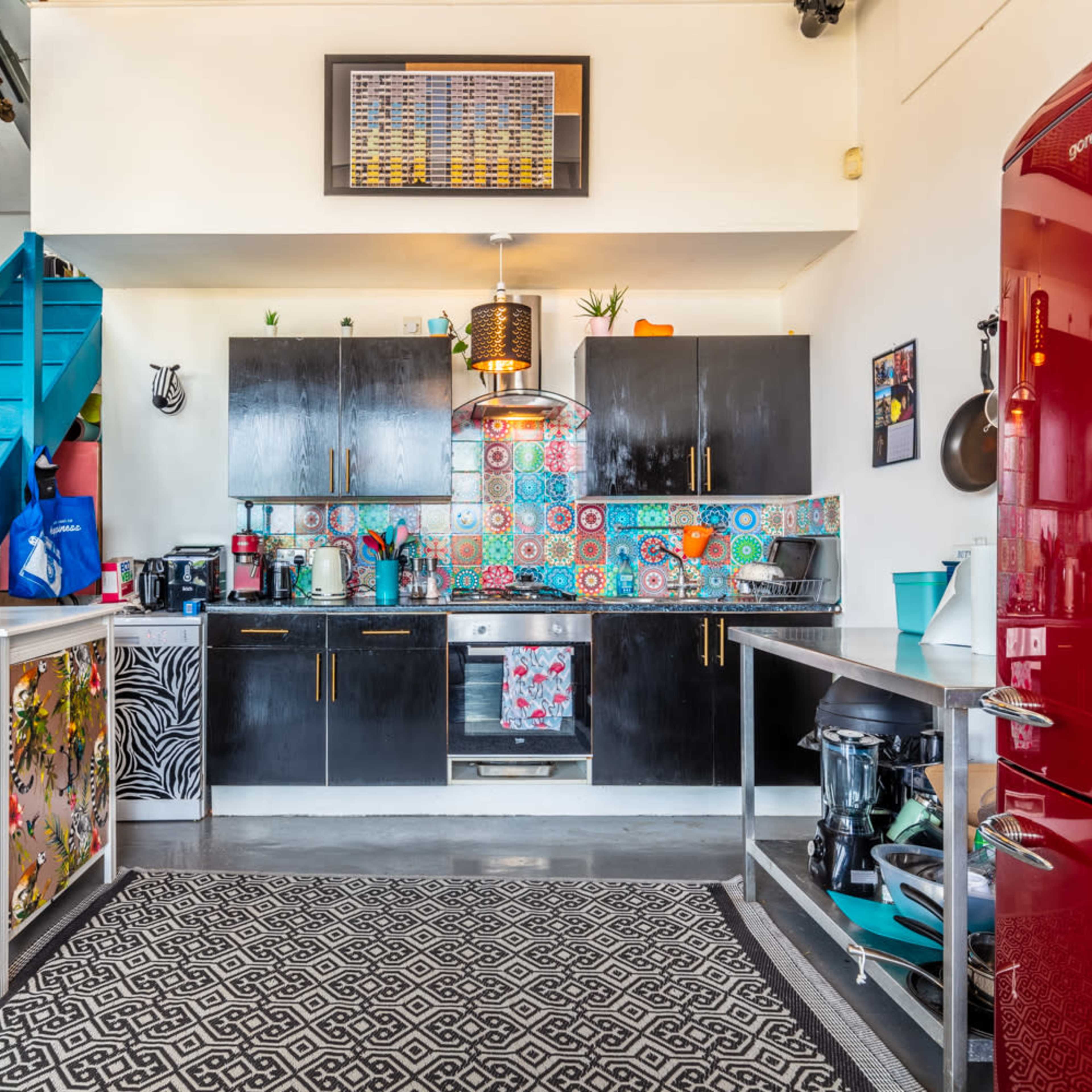 A modern kitchen featuring black cabinetry, colorful tiled backsplash, a red refrigerator, and a staircase leading to an upper level.