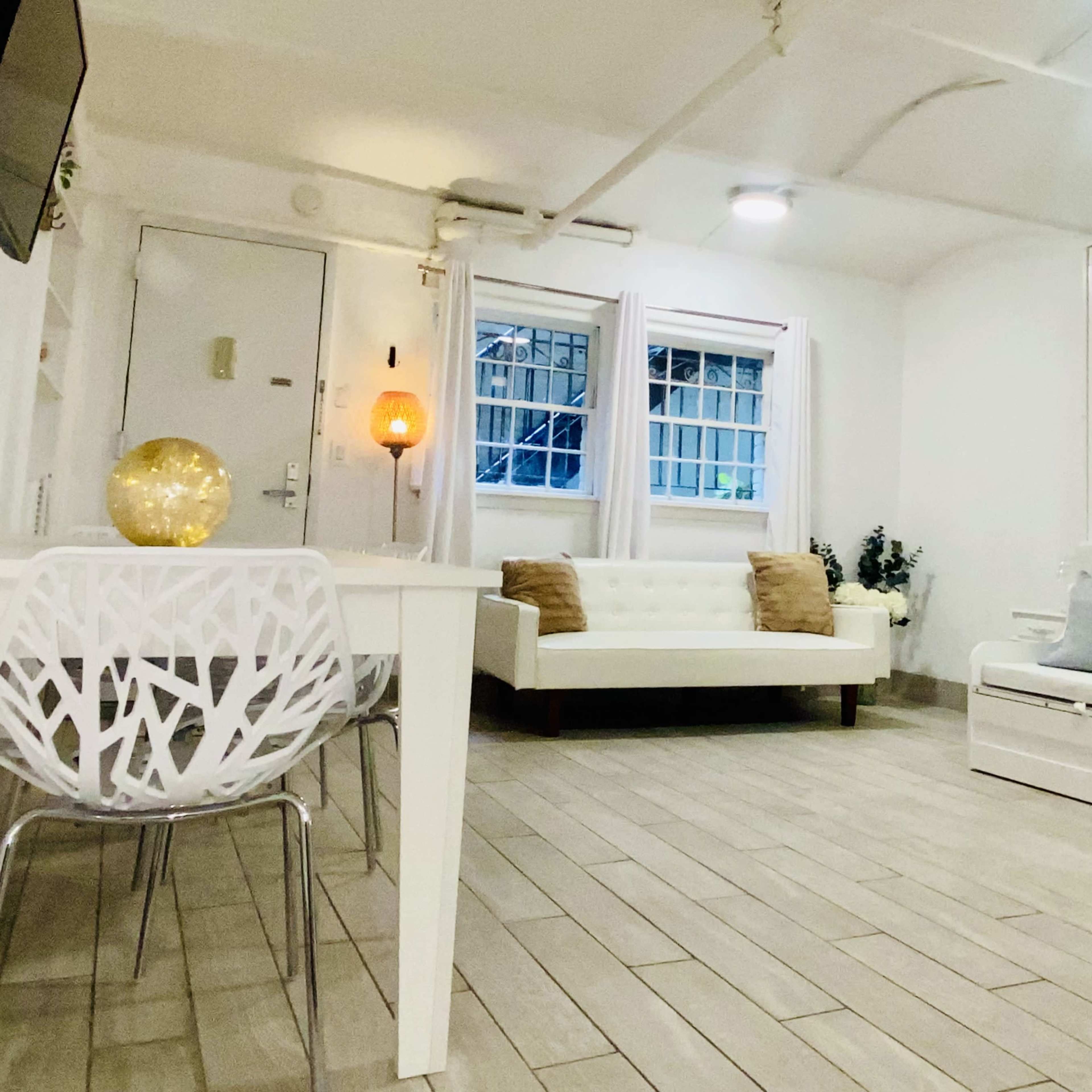 A small, modern living area with a white sofa, a dining table with decorative chairs, and light-colored tiled flooring.