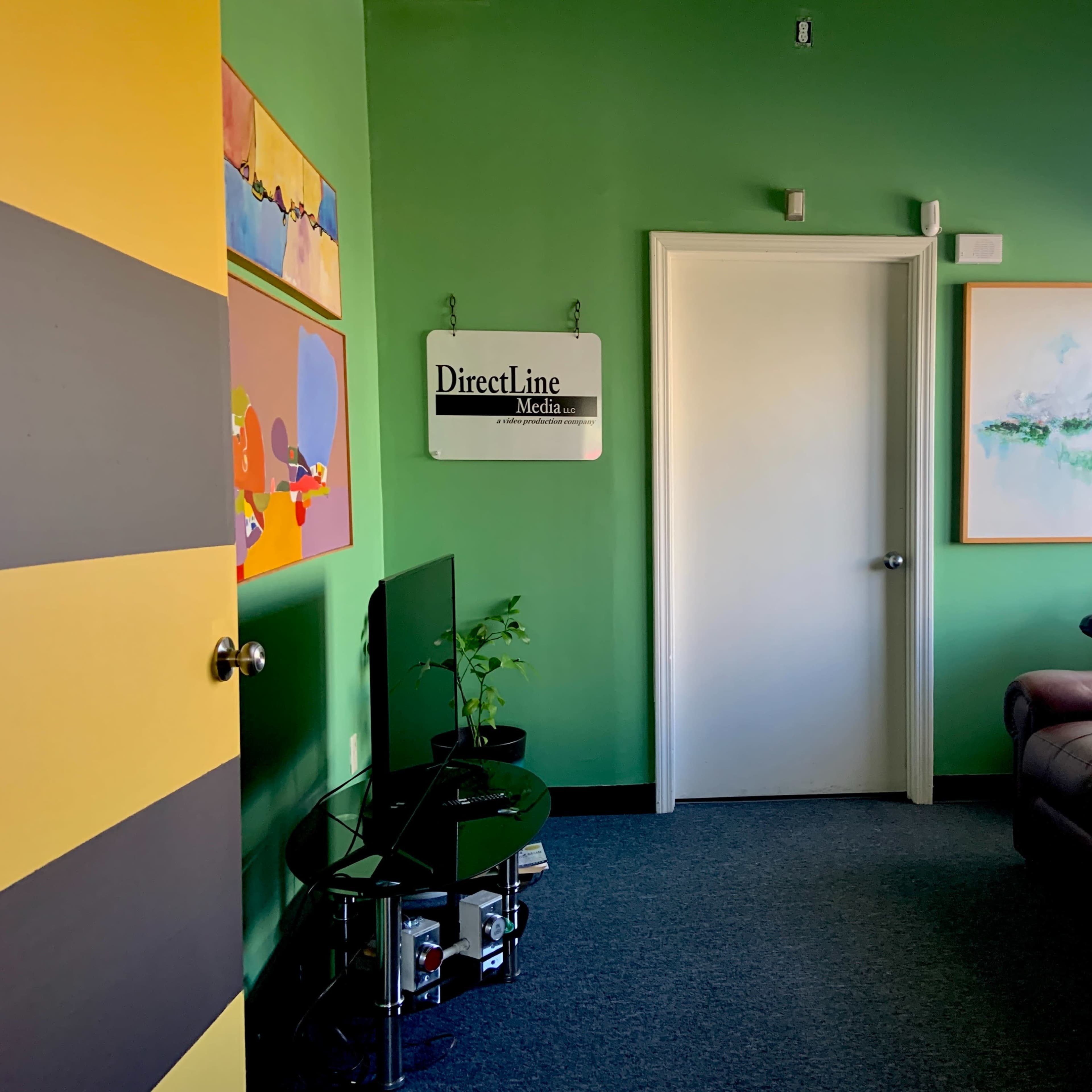 A small office room with green walls, a brown leather couch, a television on a stand, and framed artwork on the walls.