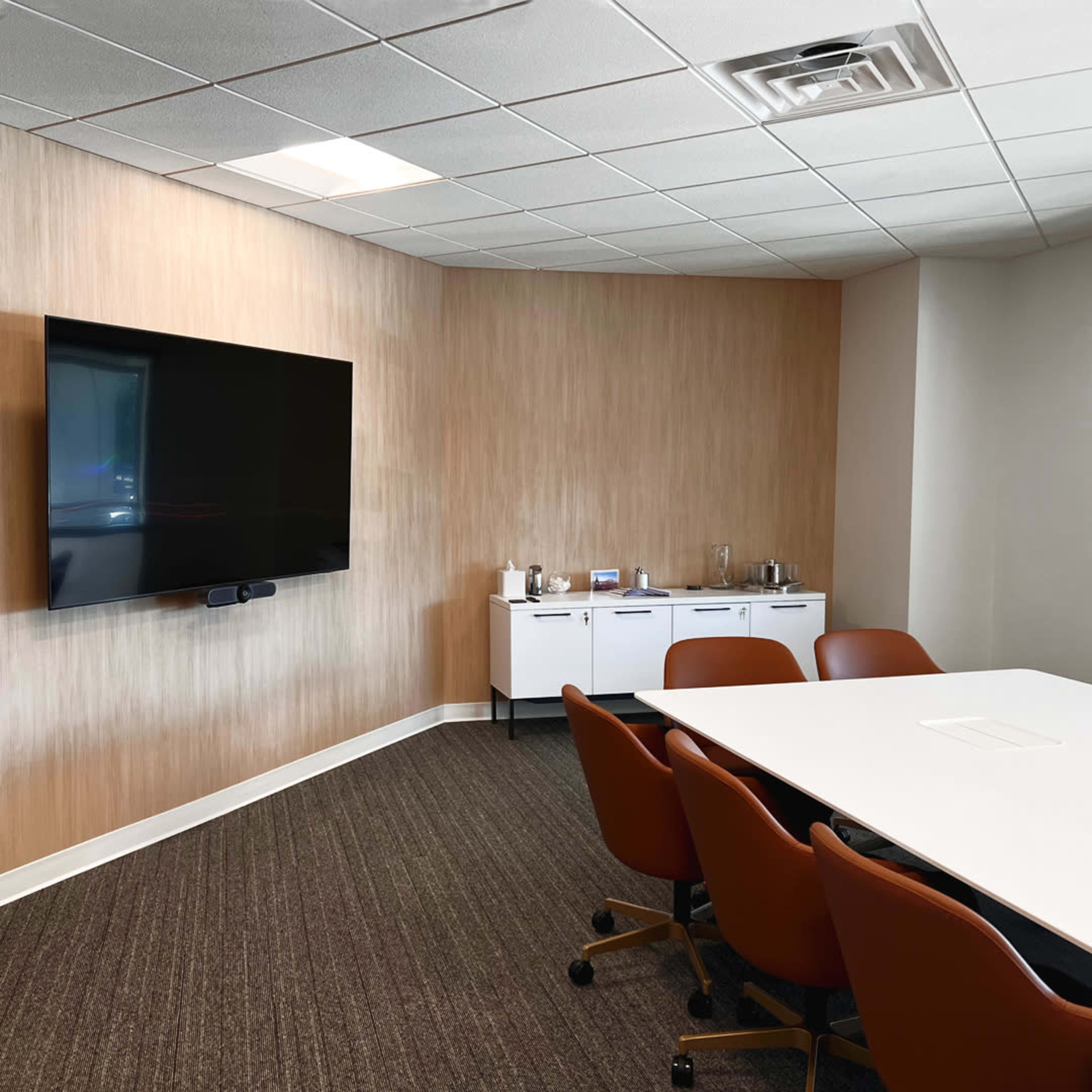 A modern meeting room with a large wall-mounted TV, a white conference table, and brown chairs.