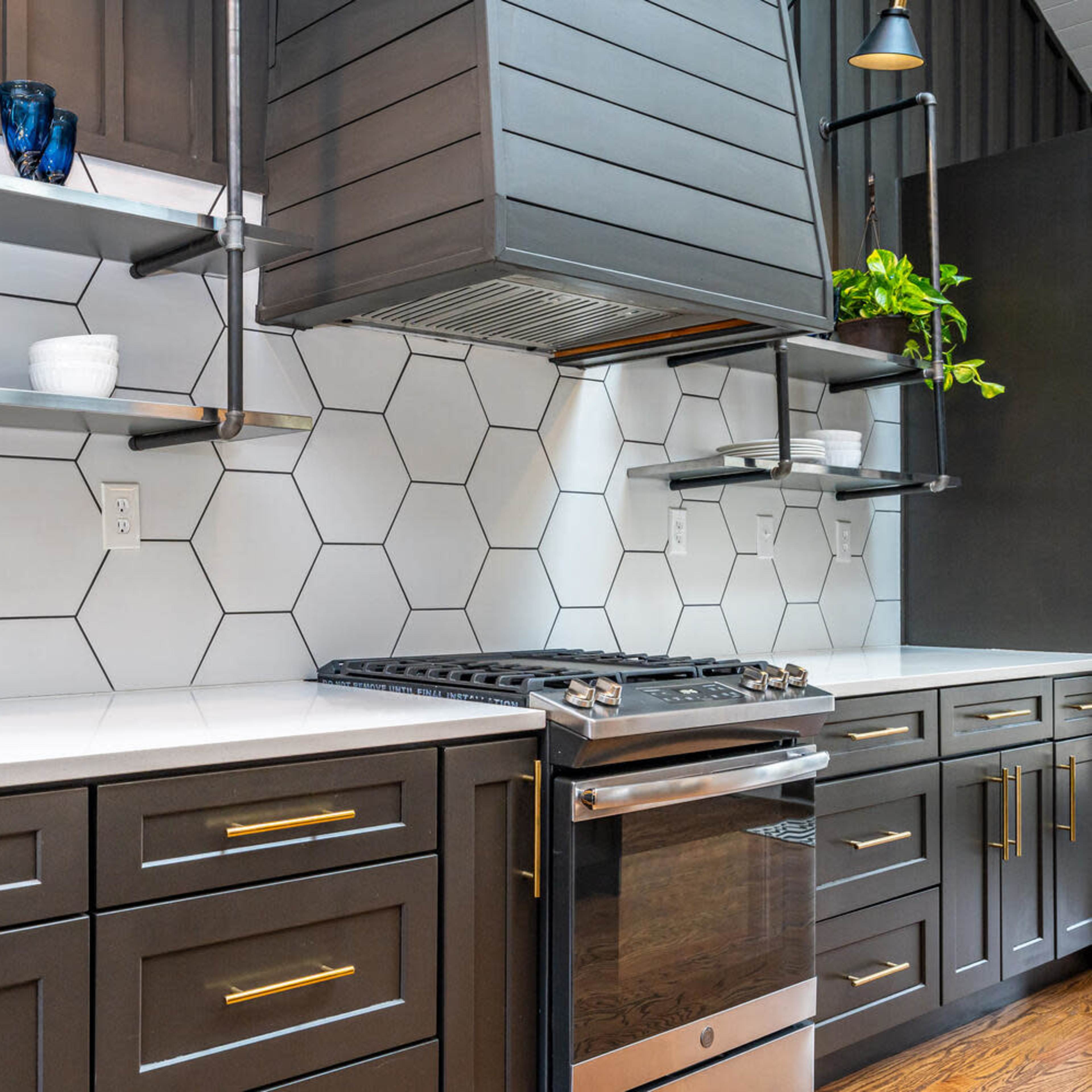 A modern kitchen with black cabinets, a stainless steel oven, and hexagonal tile backsplash, complemented by open shelving displaying plants and tableware.