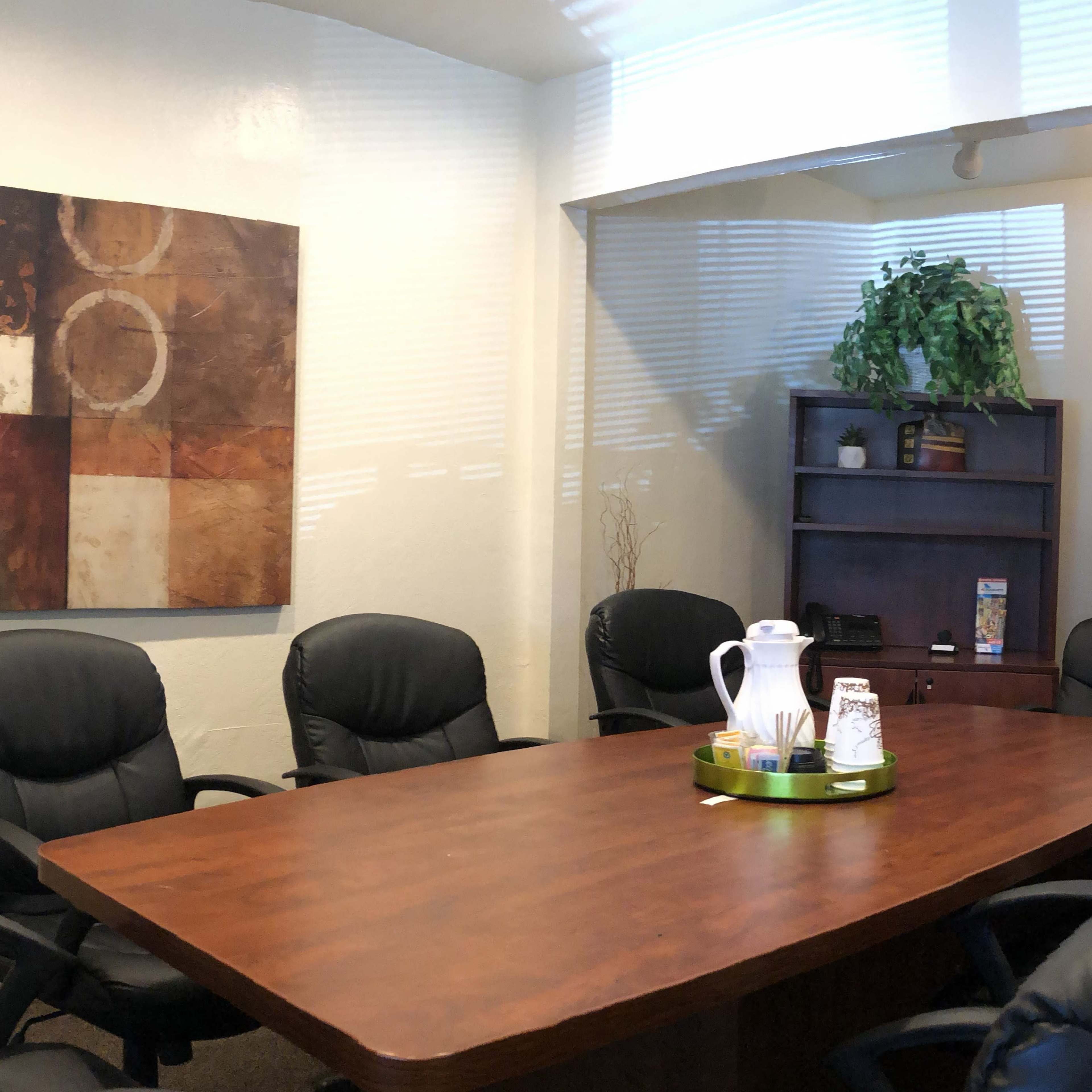 A conference room features a large wooden table surrounded by black chairs, a television, a piece of abstract art on the wall, and a small plant in the corner.