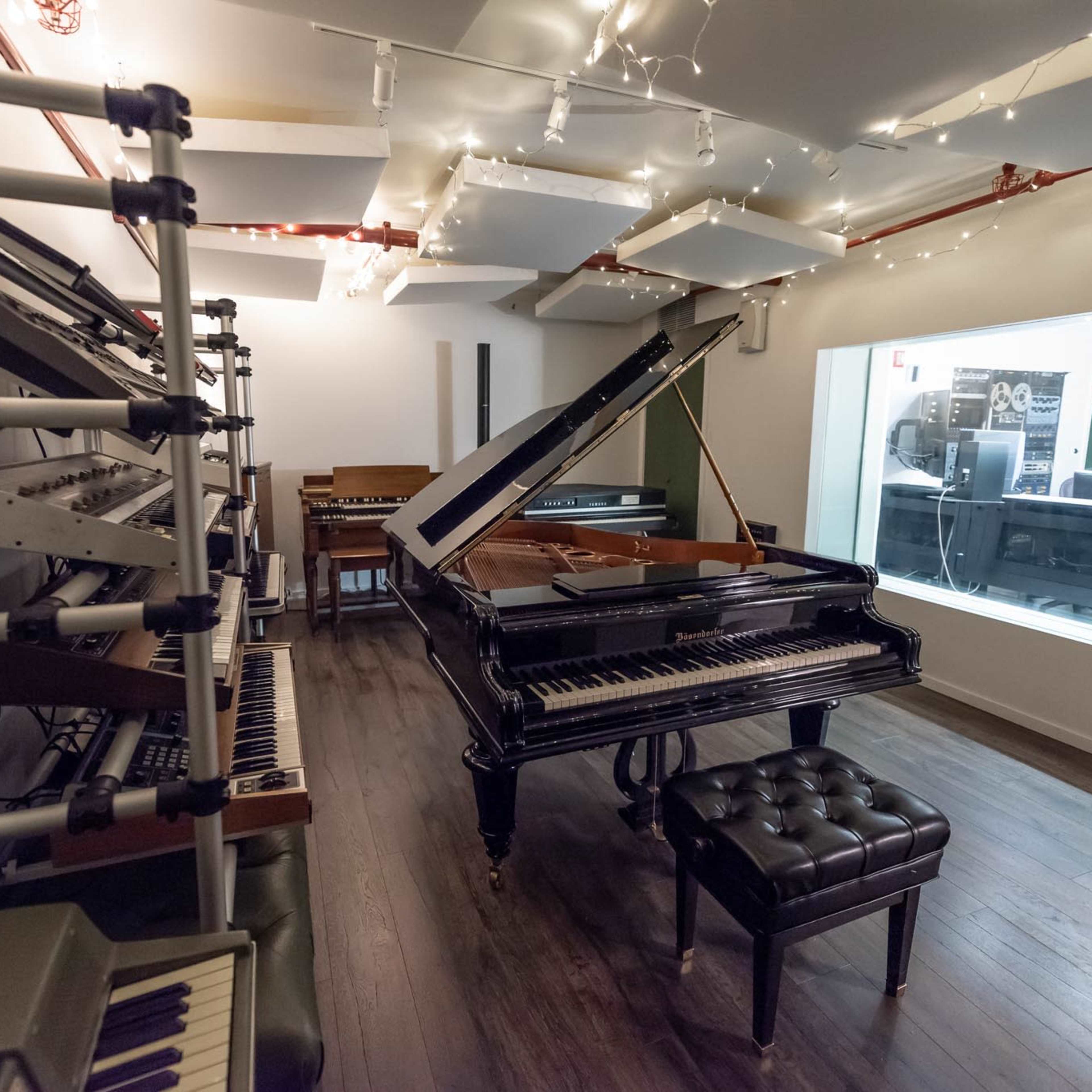 A music studio featuring a grand piano, various electronic instruments on racks, and a glass-walled control room in the background.