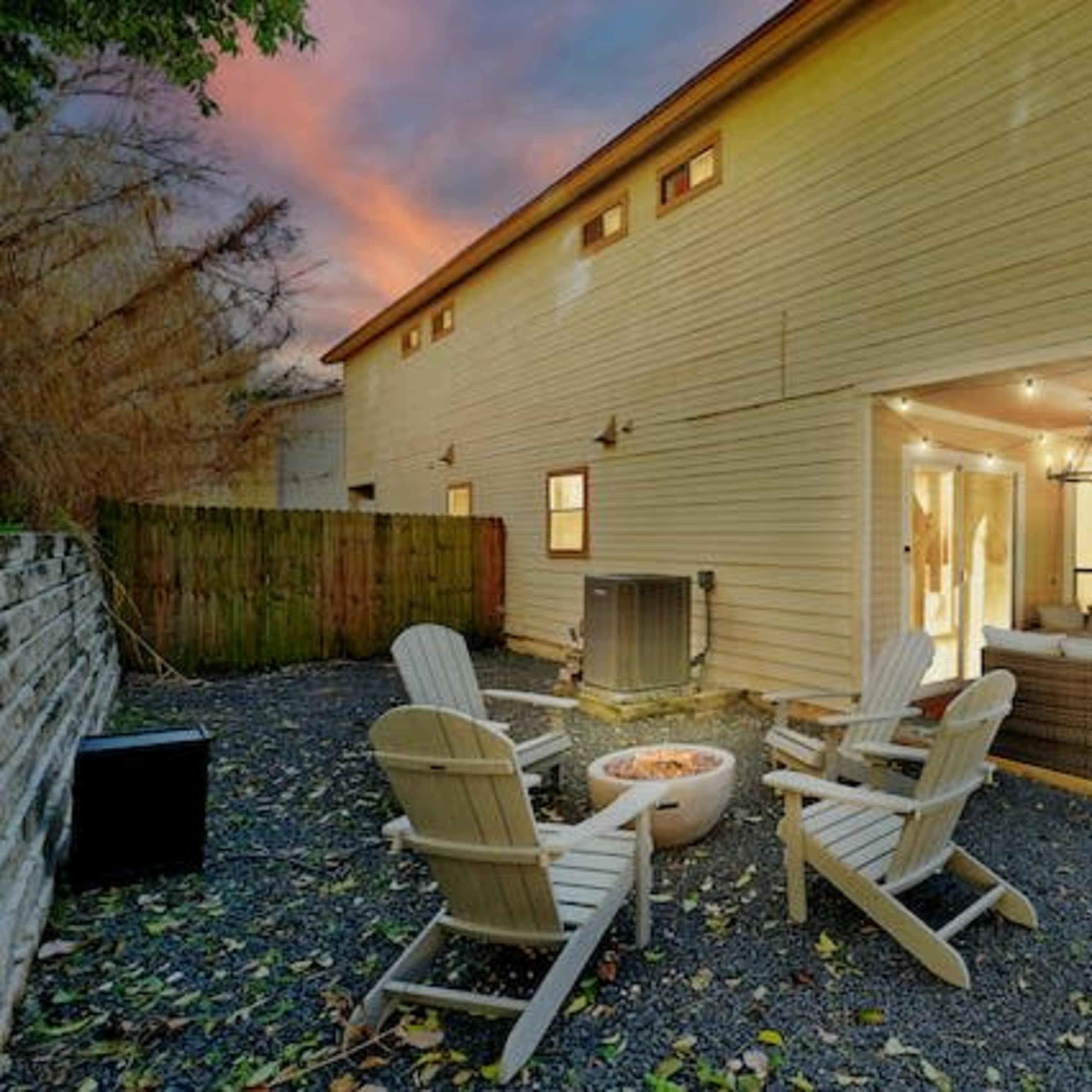 A backyard with a fire pit surrounded by four white Adirondack chairs, a patio area with seating, and a beige two-story house under a colorful sunset.