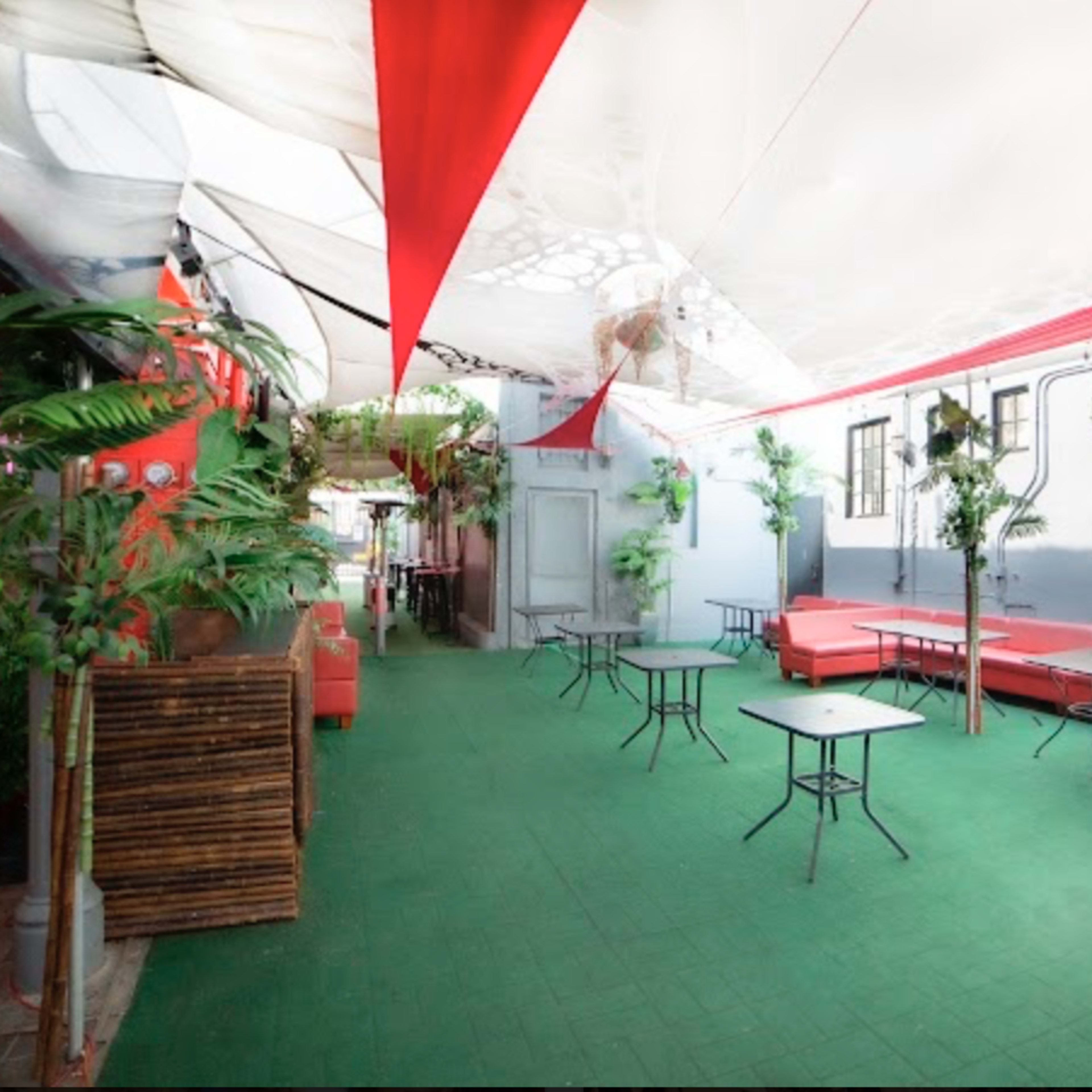 A covered outdoor area with red decor, green flooring, and small tables surrounded by potted plants.