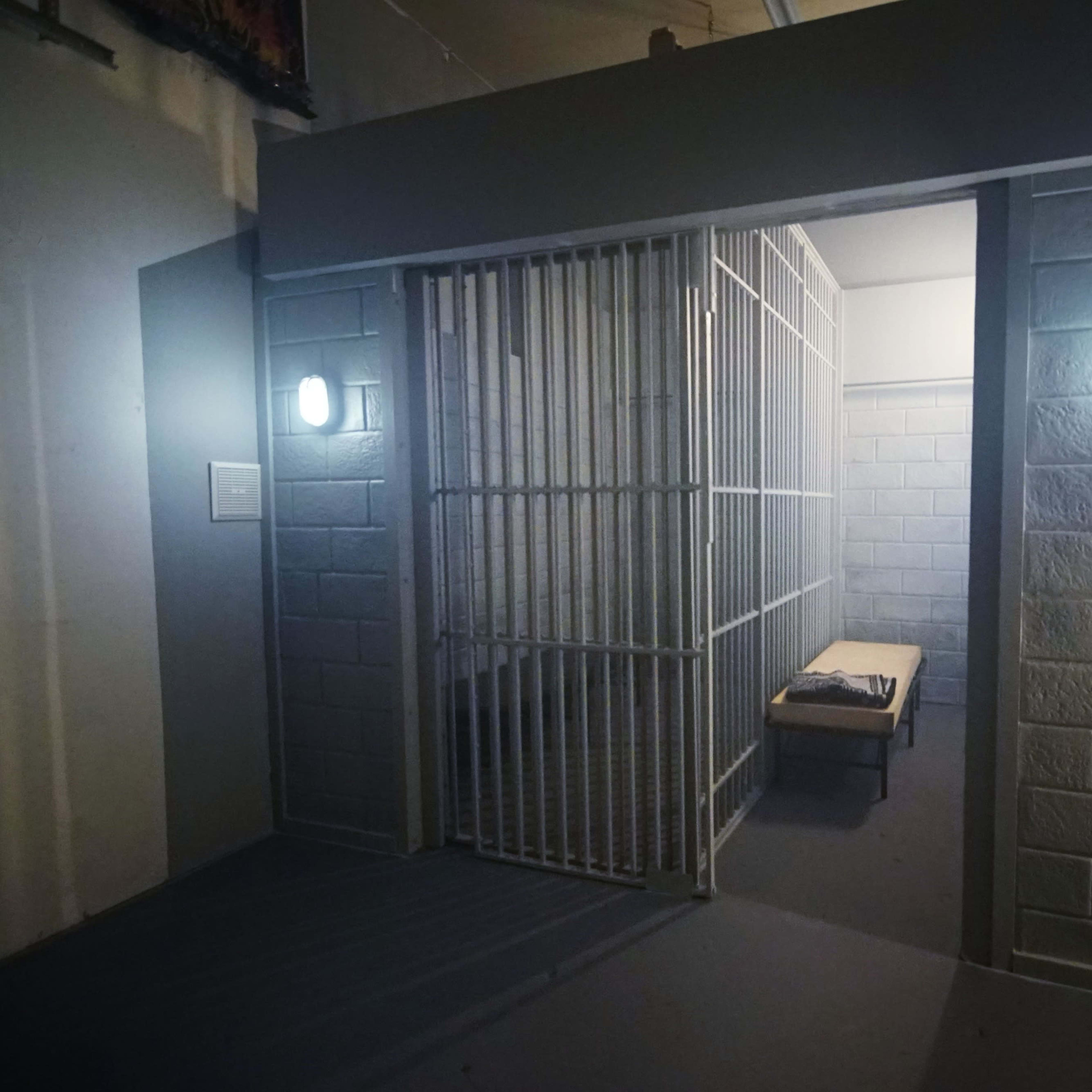 A small jail cell with a barred door and a bench inside, illuminated by two wall-mounted lights.