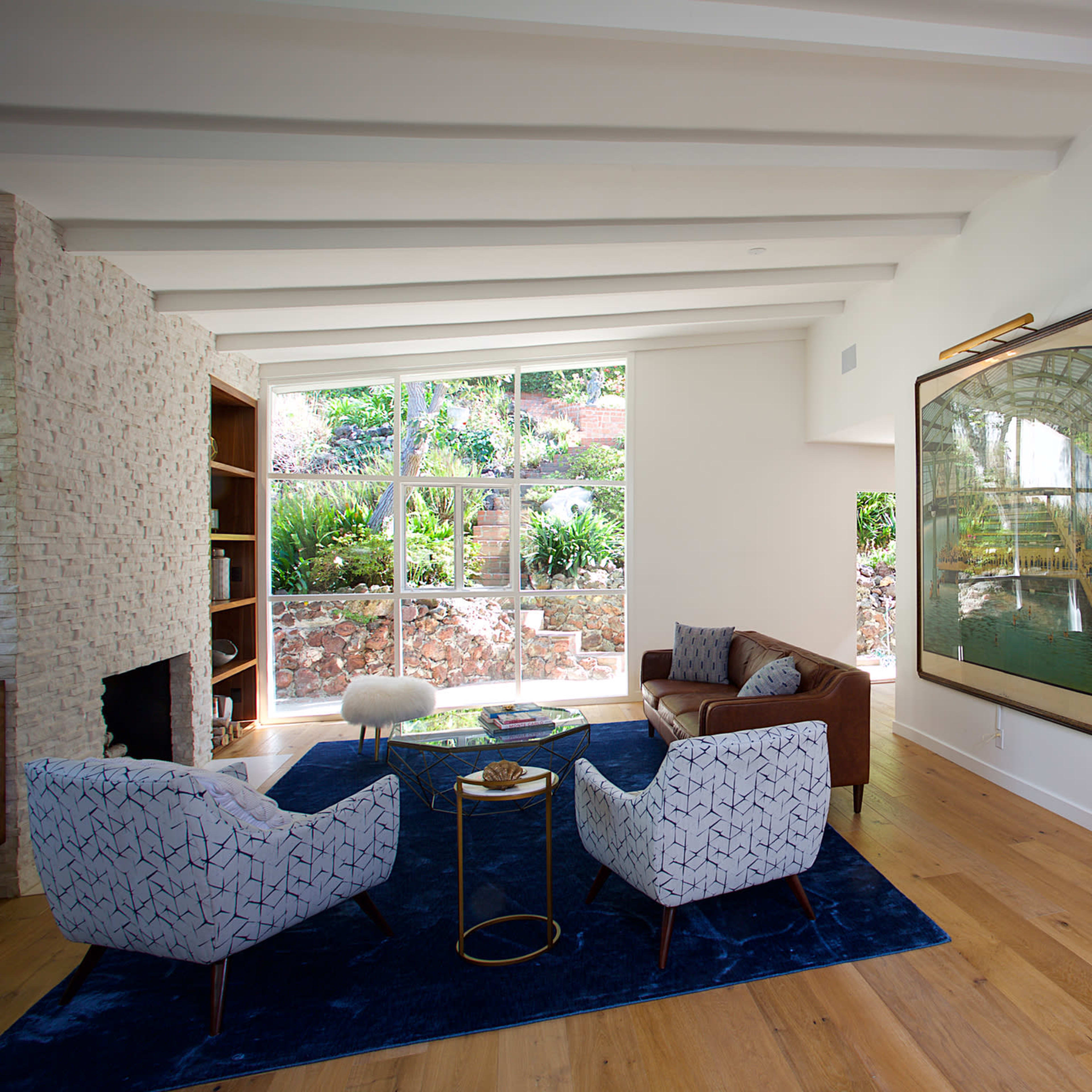 A modern living room with a blue rug, a brown sofa, two patterned armchairs, a coffee table, and large windows that provide natural light and a view of greenery outside.