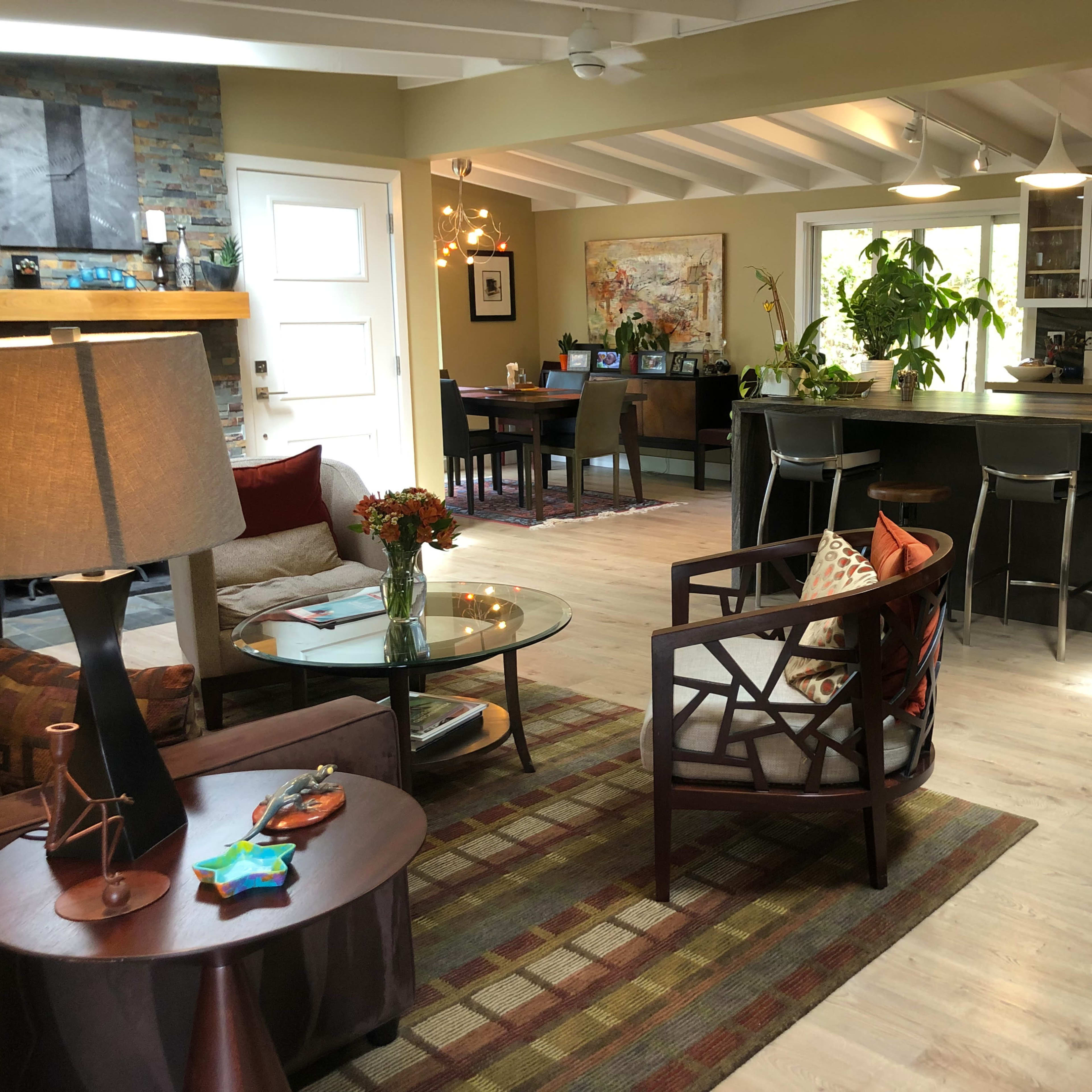 A modern open-concept living space featuring a seating area with a coffee table and chairs, a dining area in the background, and a kitchen with dark cabinetry.