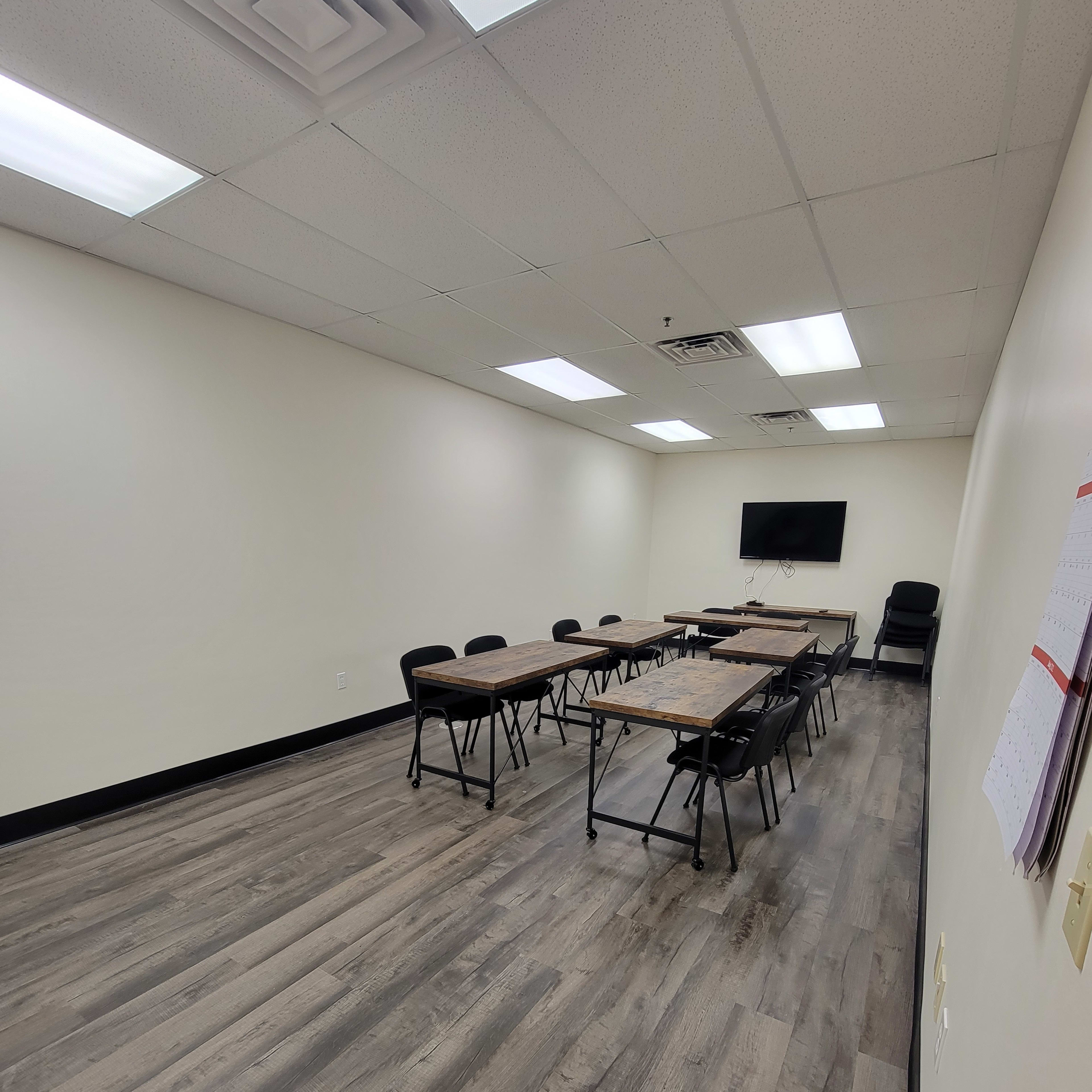 A sparsely furnished meeting room with a rectangular table, black chairs, and a wall-mounted screen.