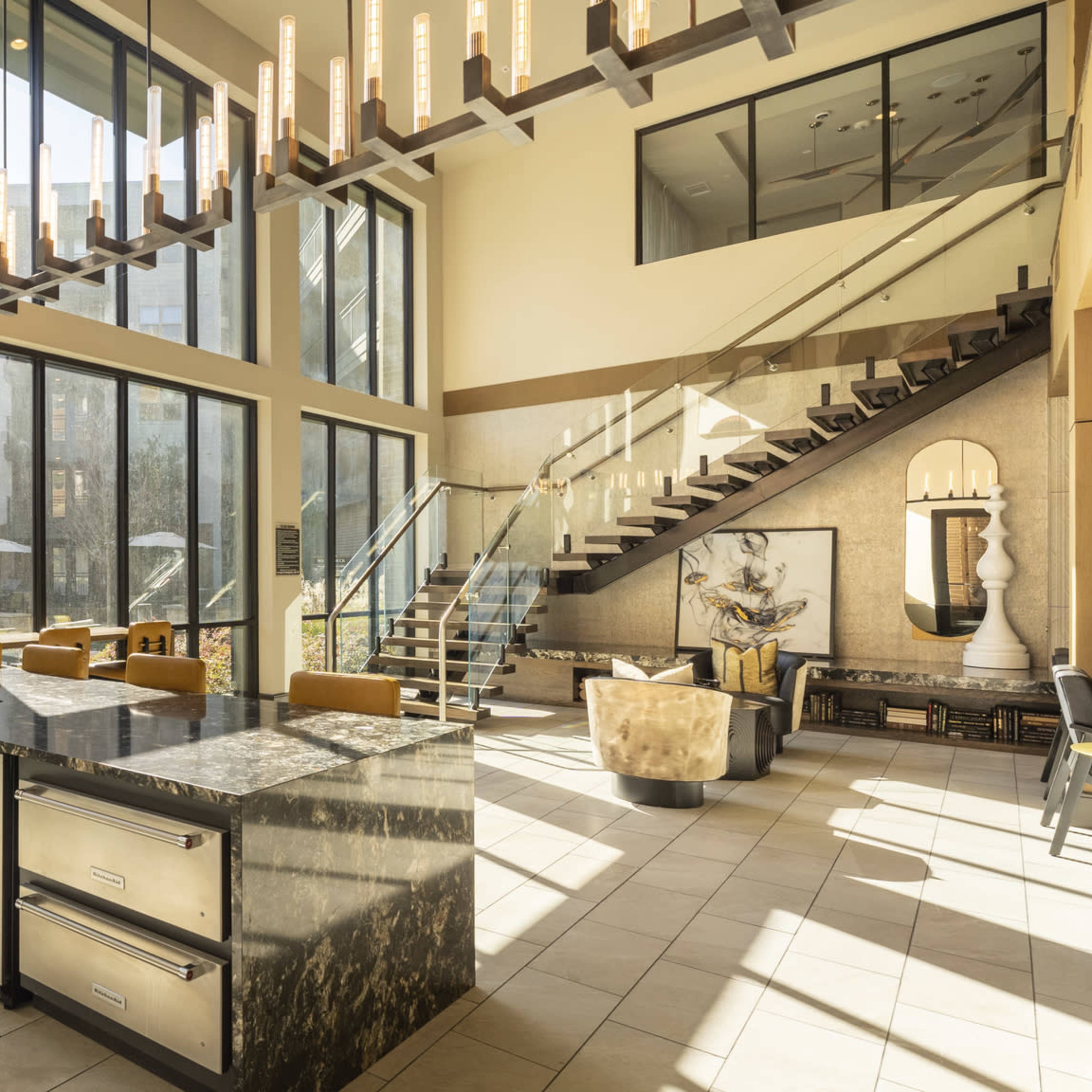 A modern open-concept living space featuring large windows, a marble kitchen island, and a staircase leading to an upper level.