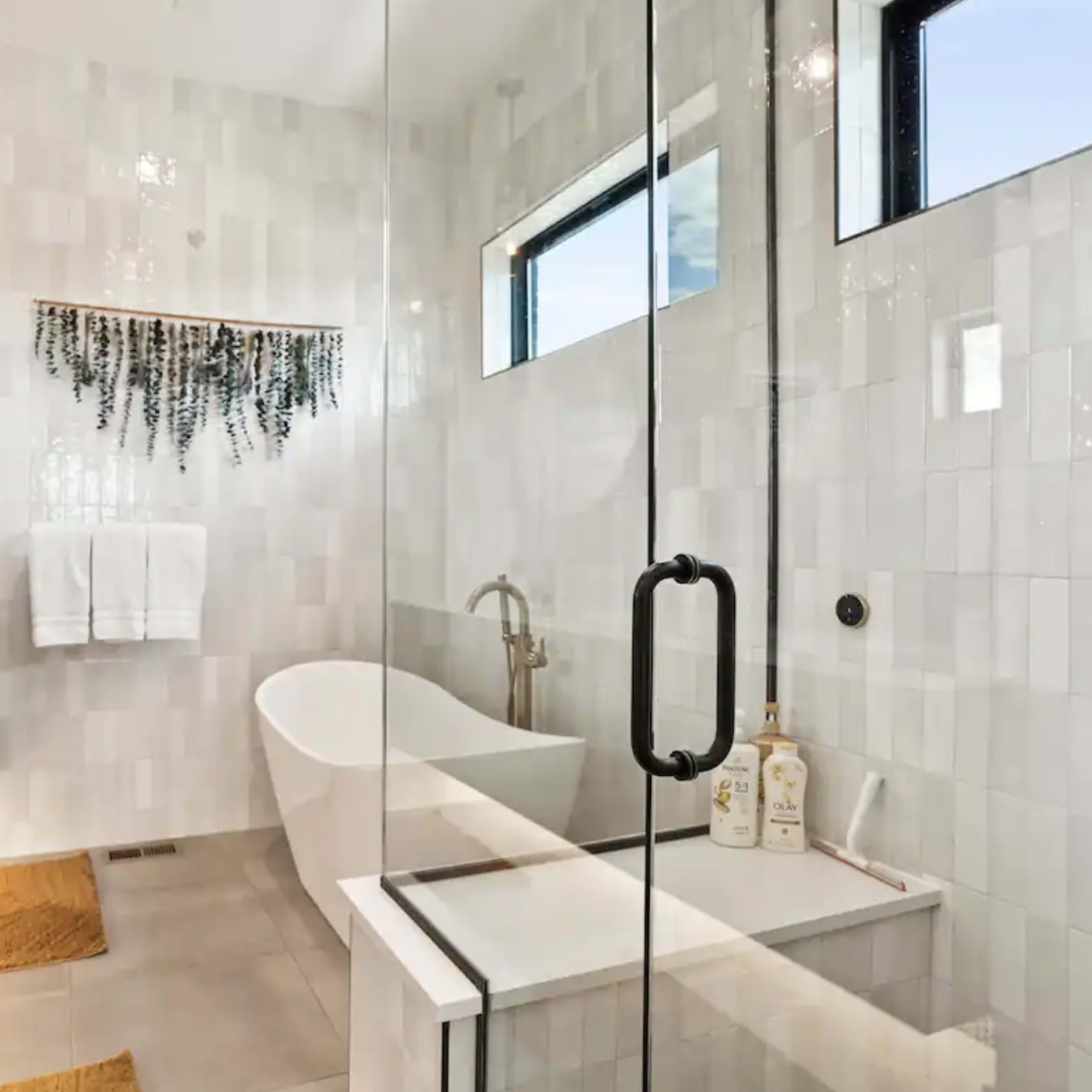 A modern bathroom featuring a glass shower, a freestanding tub, two sinks with a wooden vanity, and large wall tiles.