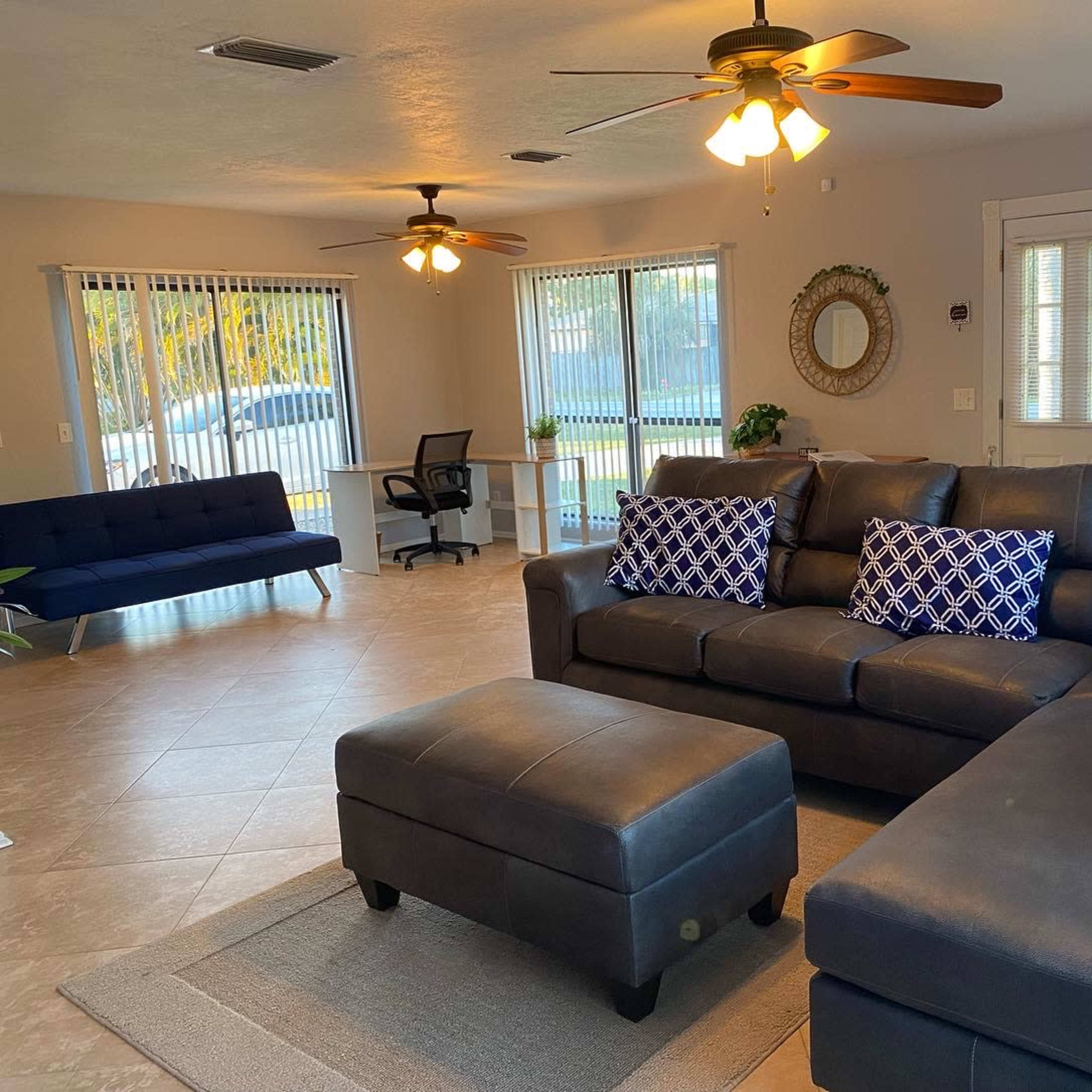 The room features a sectional sofa with blue and white pillows, a coffee table, and a desk area, along with two ceiling fans and large windows that provide natural light.